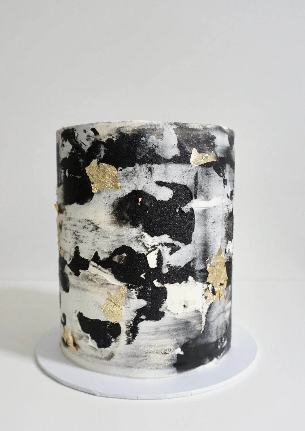 A cylindrical abstract art cake with black, white, and gold leaf accents on a white surface against a plain background.