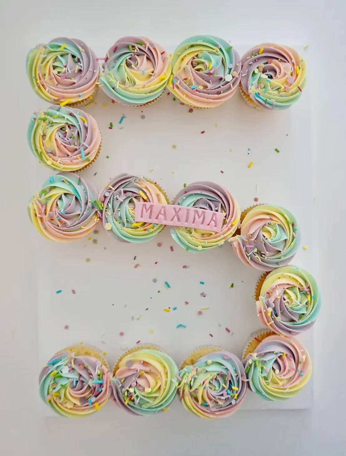 Colorful cupcakes with swirl frosting and sprinkles arranged in the shape of the letter E, with a pink fondant strip in the middle that reads 'MAXIMA'.