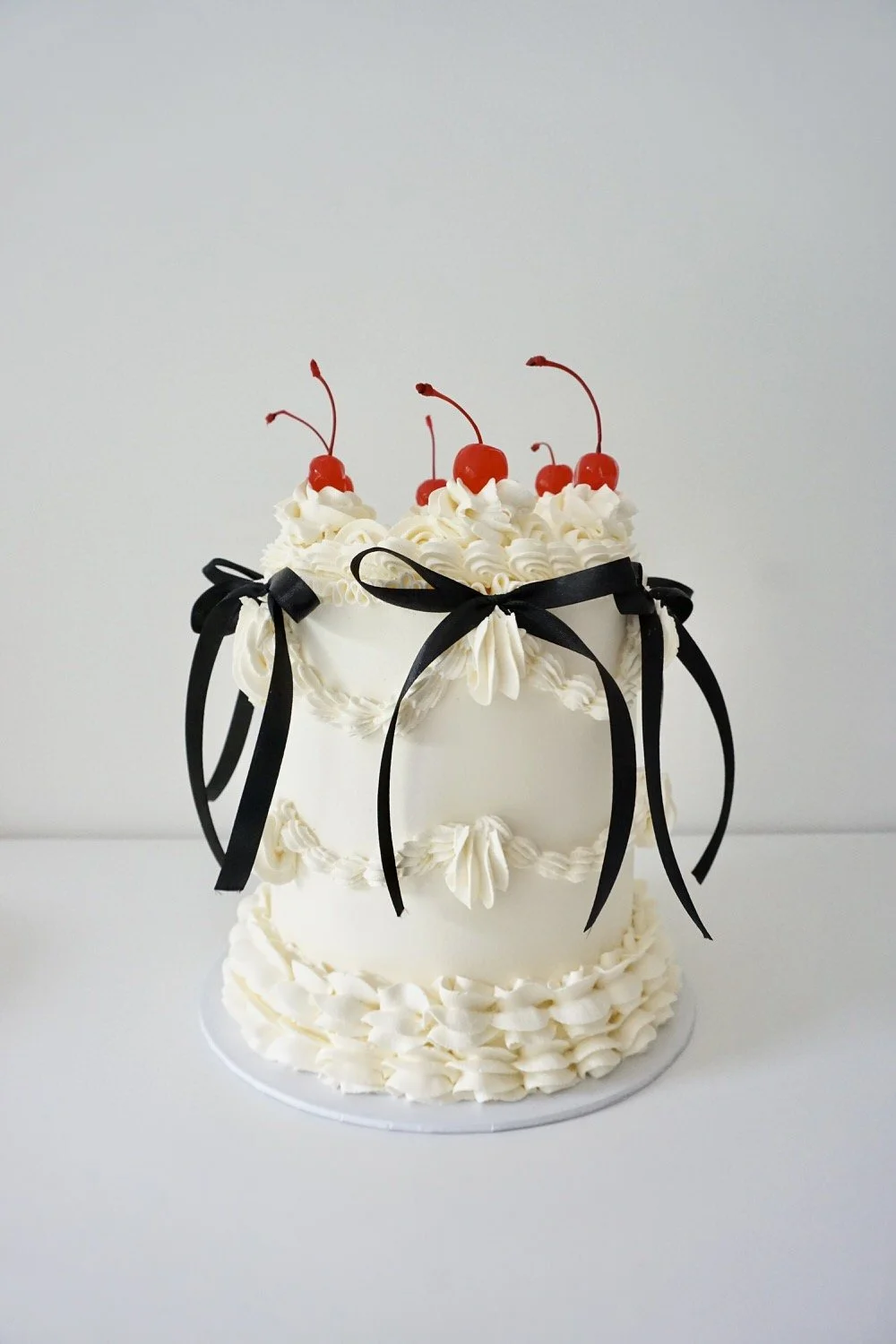 A white tiered cake decorated with whipped cream, black ribbons, and cherries on top.