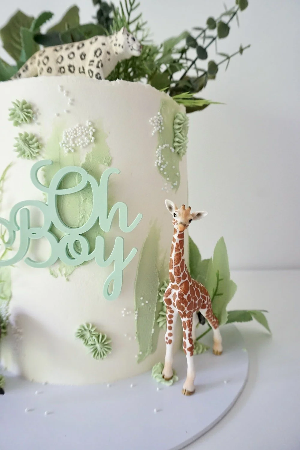 A white cake decorated with green foliage, a small plastic giraffe, and a light blue 'Oh Boy' cake topper, celebrating a baby shower or similar occasion.