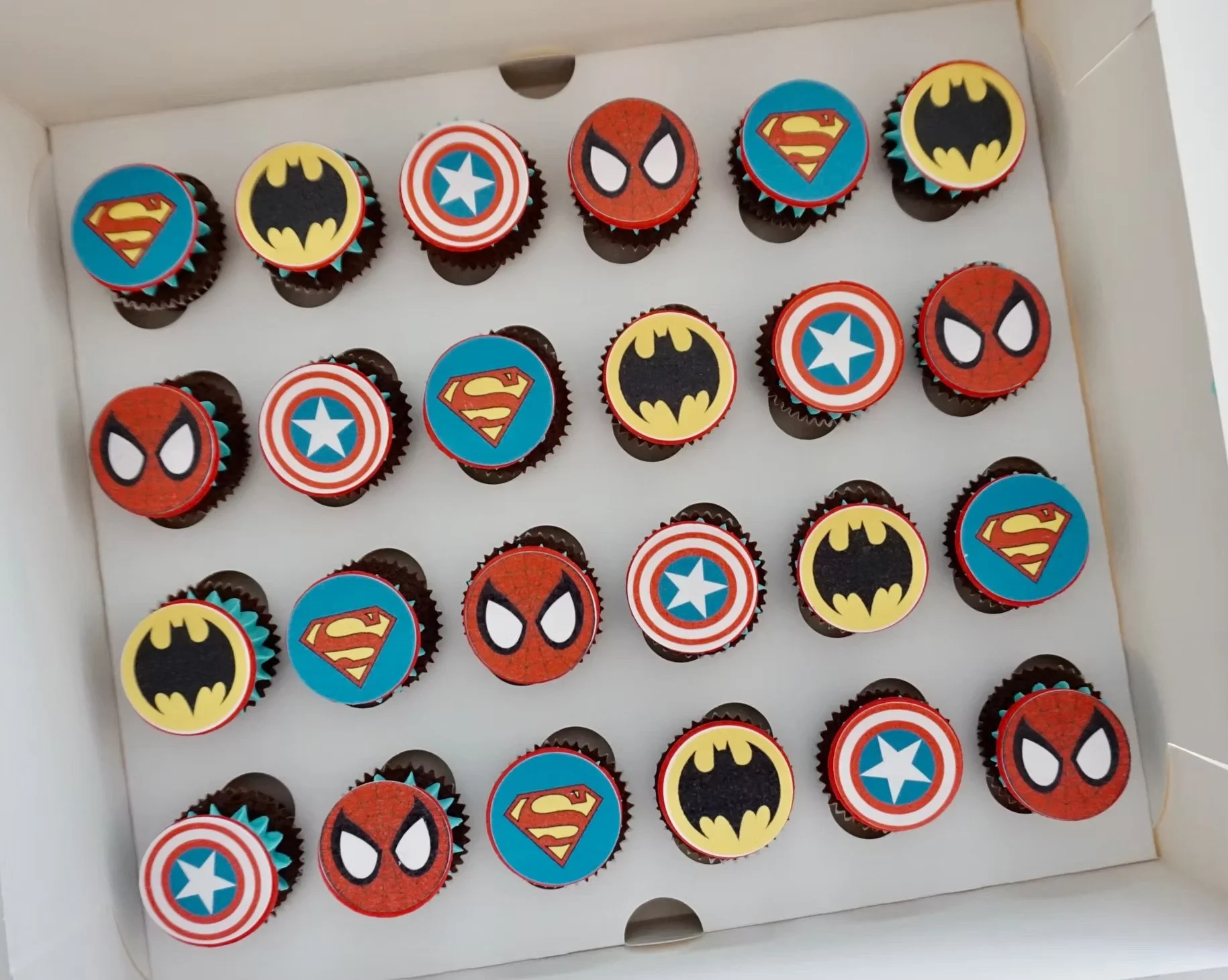 Box of cupcakes decorated with superhero themes featuring logos of Batman, Superman, Captain America, Spider-Man, and Batman symbols.