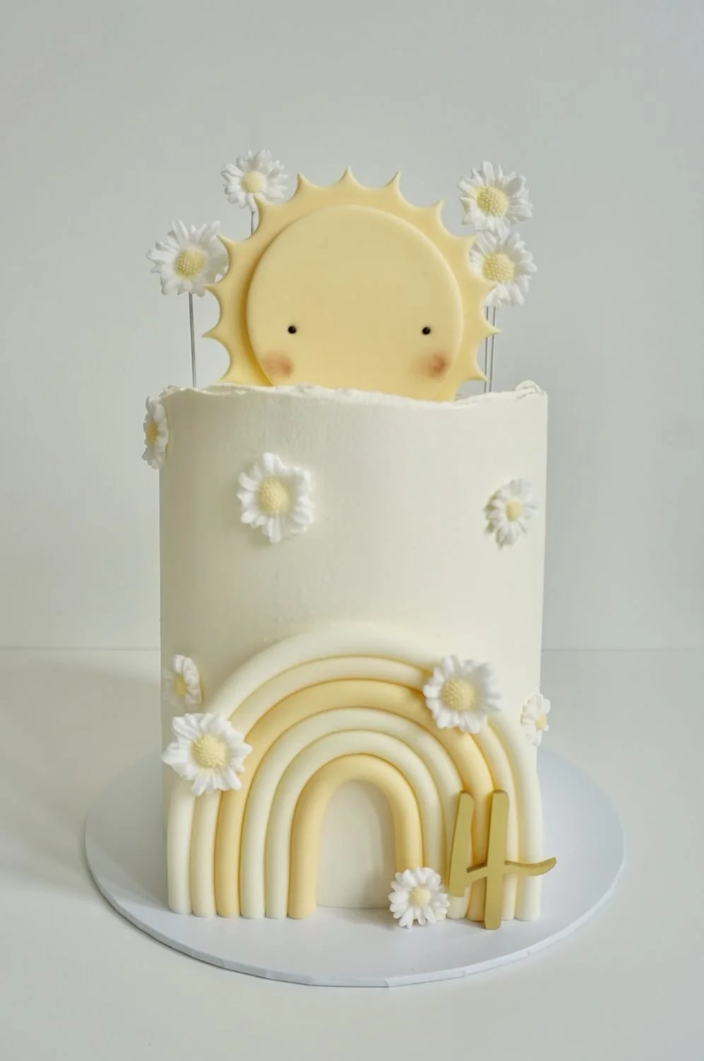White birthday cake with daisy flowers, a sun with a cute face, a rainbow, and a number 4 on the cake.