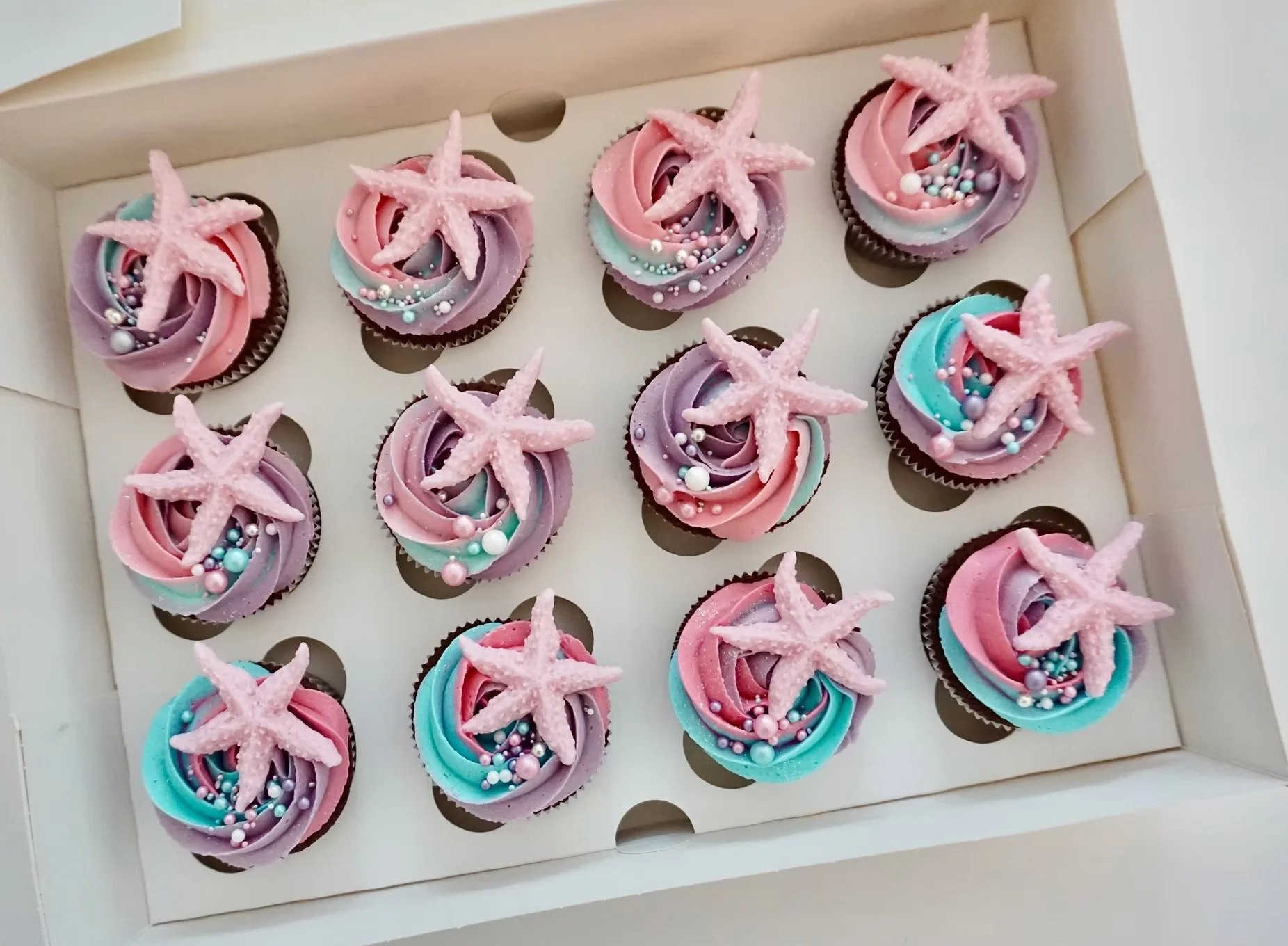 A box of 12 cupcakes decorated with colorful swirl frosting in pink, purple, and blue, topped with pink starfish-shaped decorations and small round pearl-like sprinkles.