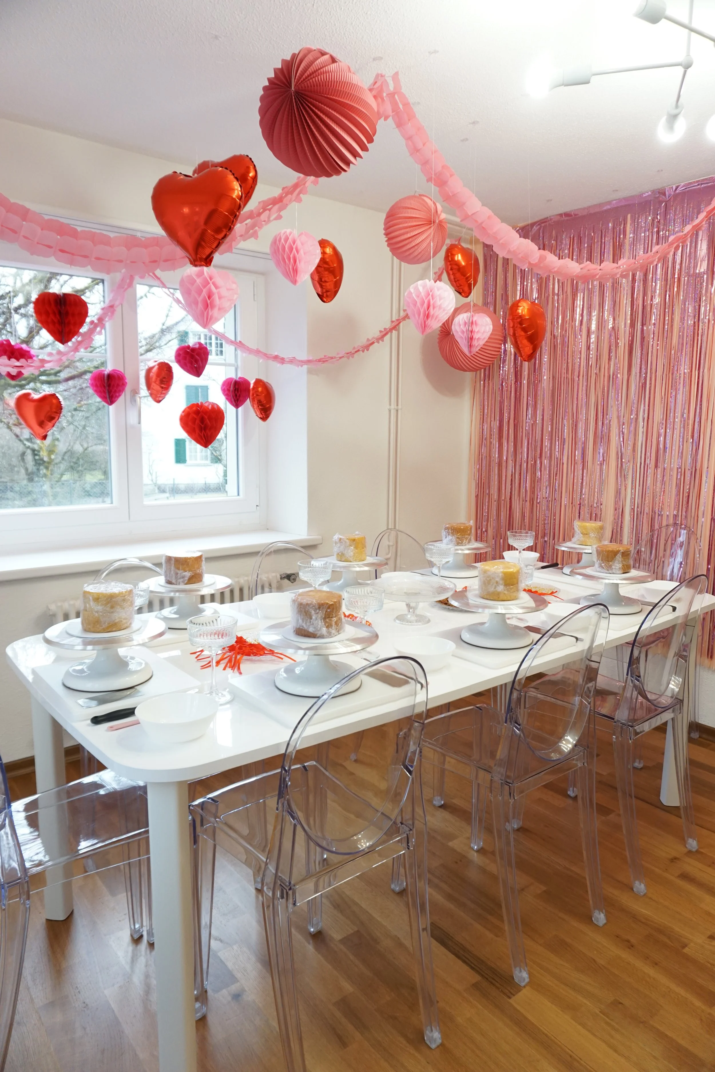 Valentine's Day decorated dining table with pink and red heart-shaped balloons and paper decorations, set with plates, bowls, glasses, knives, and forks, near a window with pink curtains.