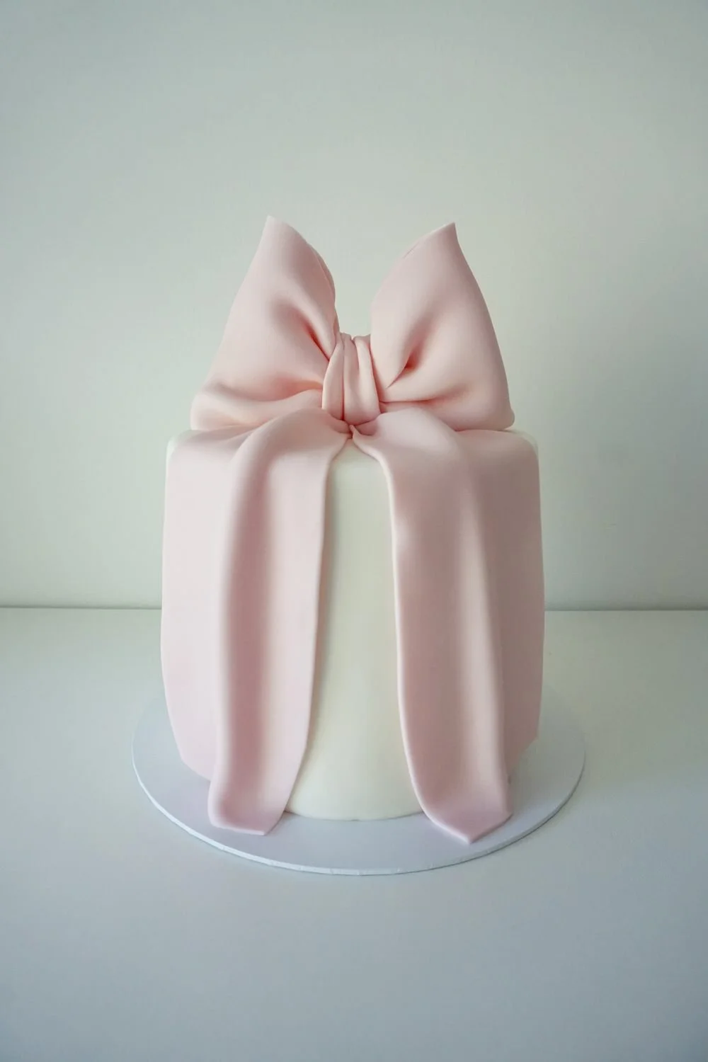 A cake decorated with a large pink fondant bow on top, with long tails cascading down the sides, set against a plain background.