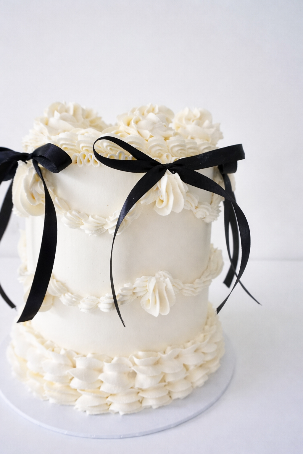 A three-tier white wedding cake decorated with white buttercream piping and topped with black ribbons tied in bows.