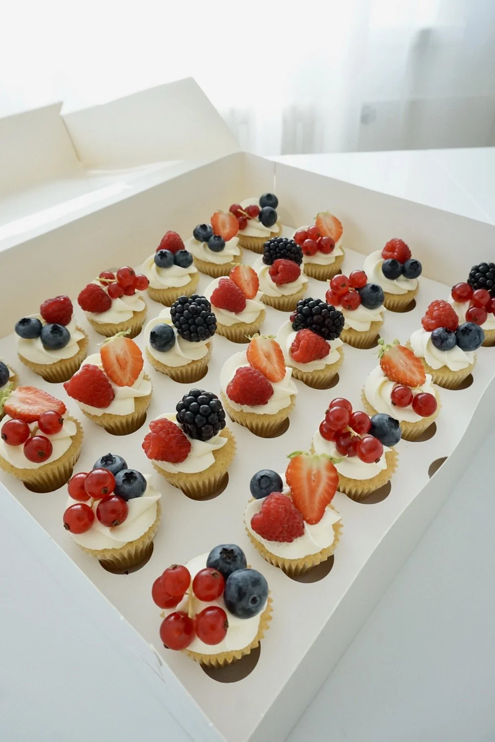 A box of assorted mini fruit-topped cupcakes with blueberries, strawberries, raspberries, blackberries, and red currants.
