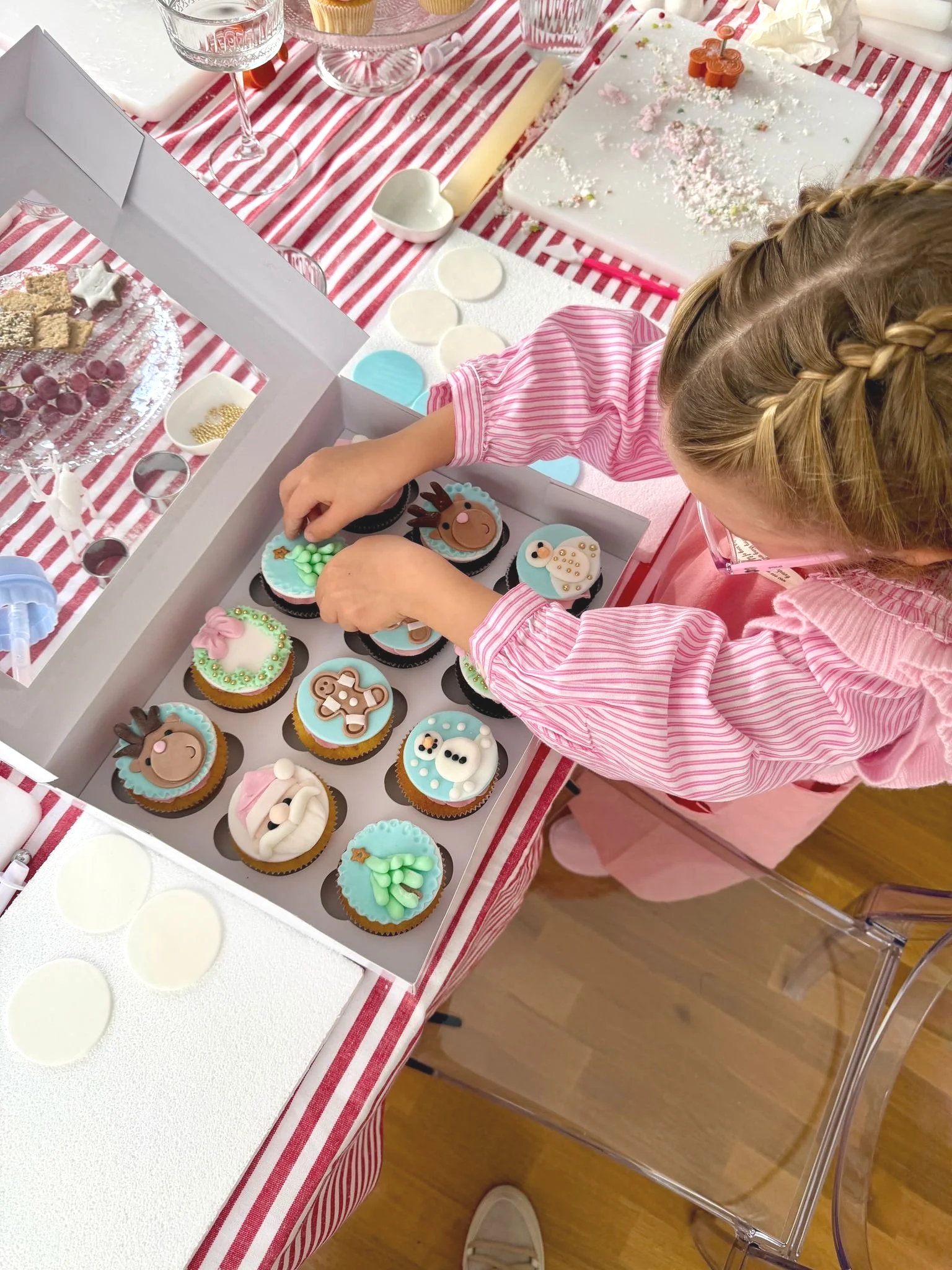 A young girl in a pink striped dress decorating cupcakes with holiday-themed frosting, including reindeer, snowmen, and other festive designs, on a table covered with a red and white striped tablecloth.