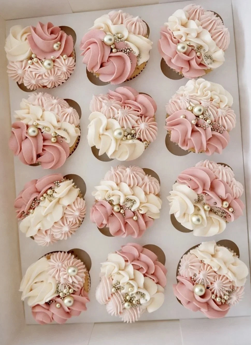 A box of twelve cupcakes decorated with pink and white frosting, pearls, and silver beads.