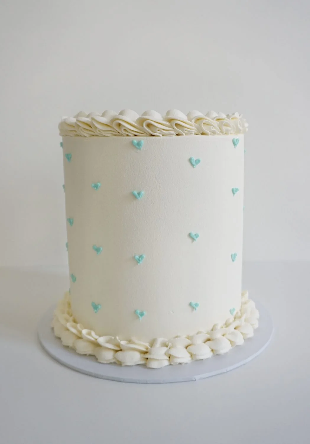 A tall, white frosted birthday cake with small blue heart decorations, piped white border at the top and bottom, and a border of white frosting shells at the base, set on a gray cake board.