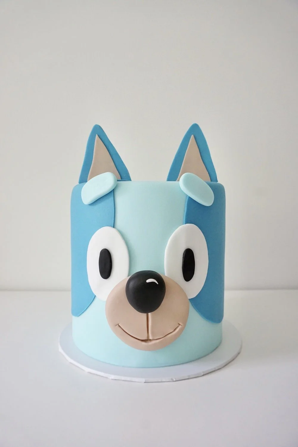 Cake shaped like a cartoon dog face with blue ears, large eyes, black nose, beige mouth, and blue and beige fur, on a white surface against a plain background.