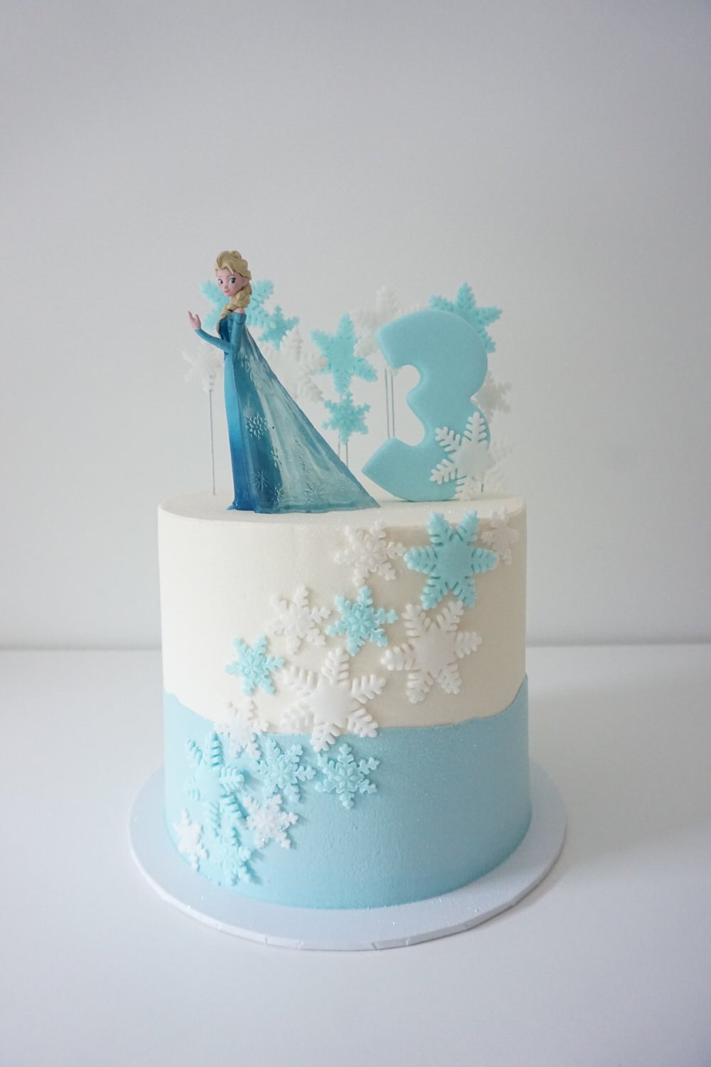 White and blue birthday cake with snowflake decorations, an Elsa figurine from Frozen, and a large blue number 3