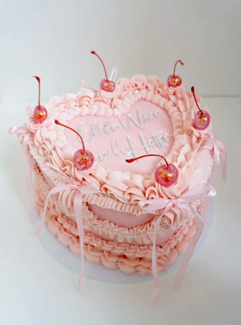 A pink birthday cake decorated with cherries, pink icing, bows, and glitter, with the message "Happy Mother’s Day" written on top.
