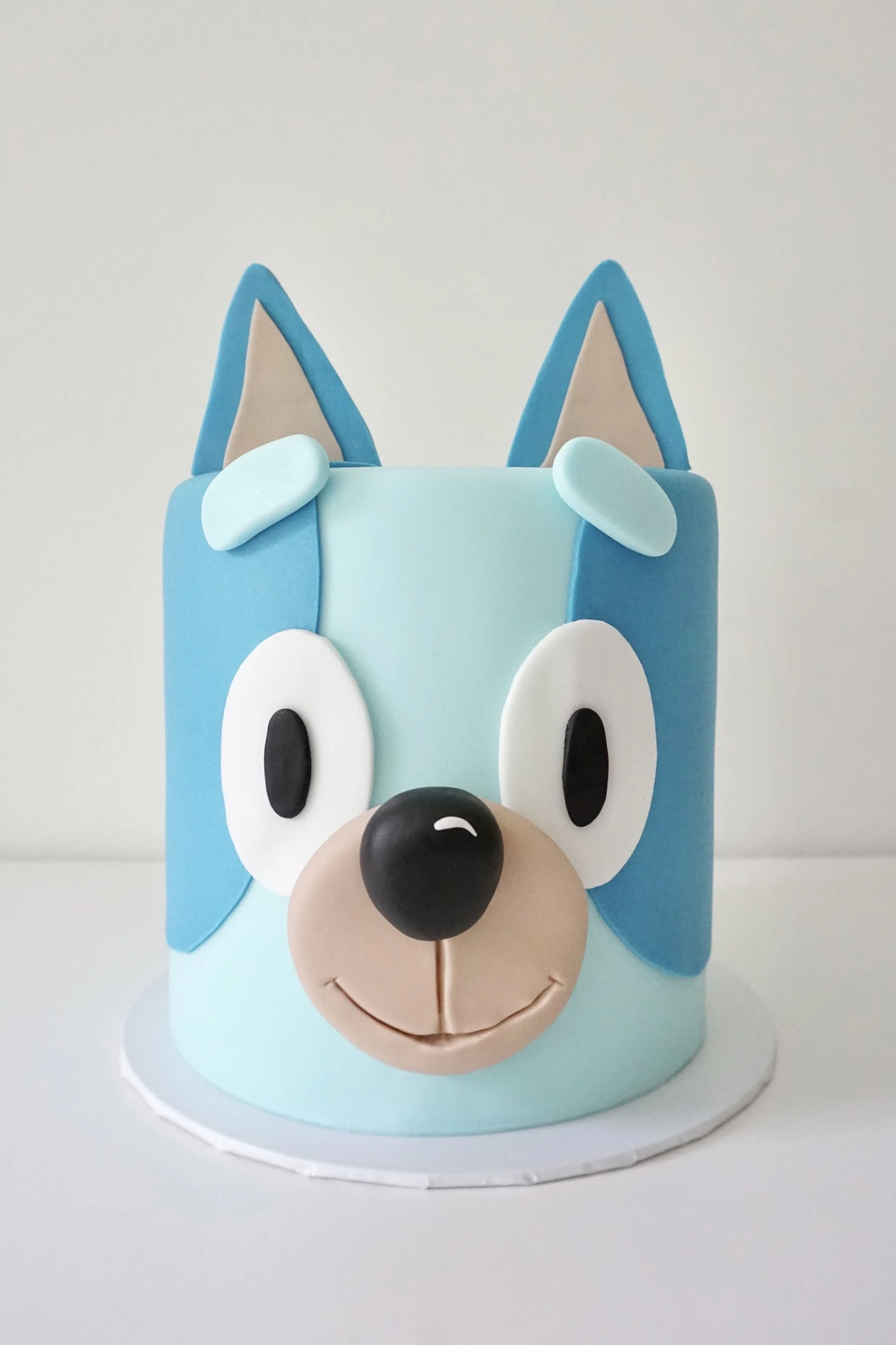 Light blue birthday cake decorated to look like a cartoon dog with blue ears, white eyes, black pupils, a black nose, and a smiling mouth, set on a small white base against a plain background.