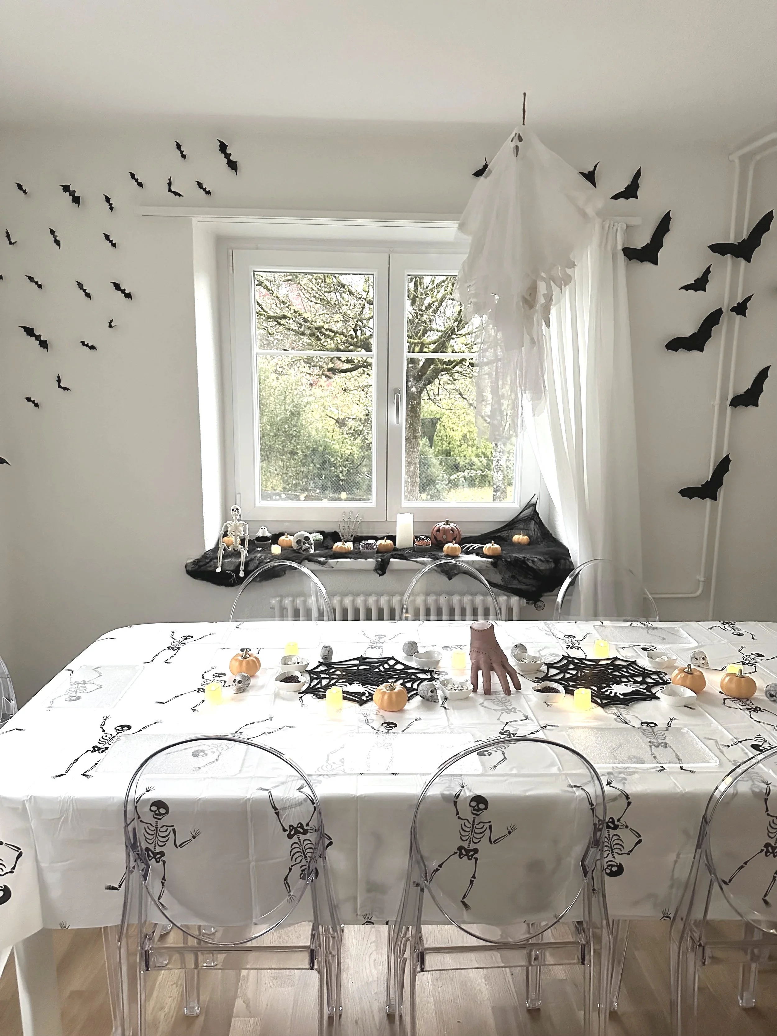 Halloween themed dining room with a table covered in a white skeleton pattern tablecloth, decorated with small pumpkins, skulls, candles, and cobweb centerpieces. The wall has black bats decorations, and a ghostly figure hangs from the ceiling near a window with a view of trees outside.