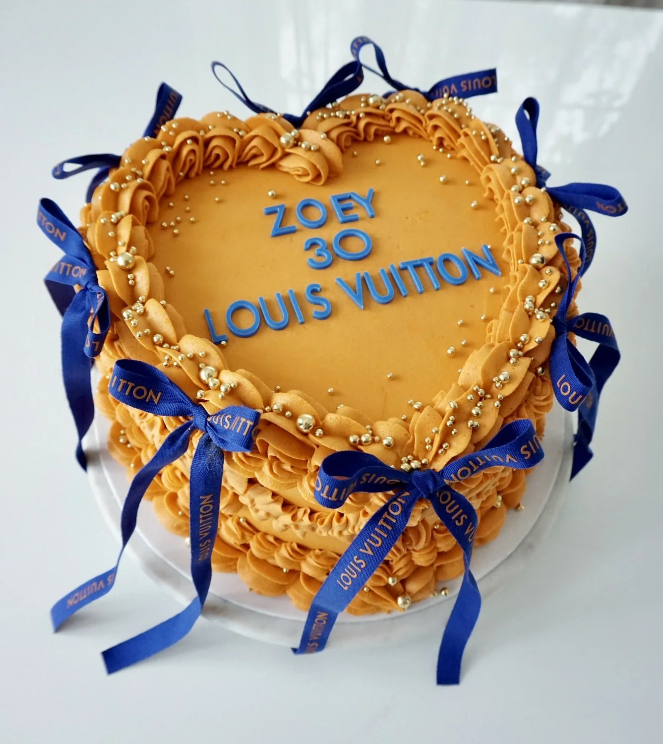 Heart-shaped birthday cake decorated with orange frosting, gold and silver edible beads, and blue ribbons with 'Louis Vuitton' written on them. The cake has blue lettering that reads, 'Zoey 30 Louis Vuitton' in the center.