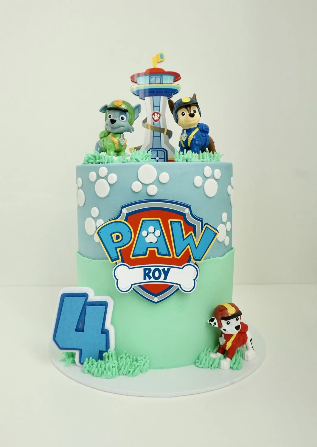 Paw Patrol themed birthday cake with two tiers, decorated with paw print patterns, a Paw Patrol logo, and figurines of Chase, Marshall, and Rubble on top, against a plain white background.