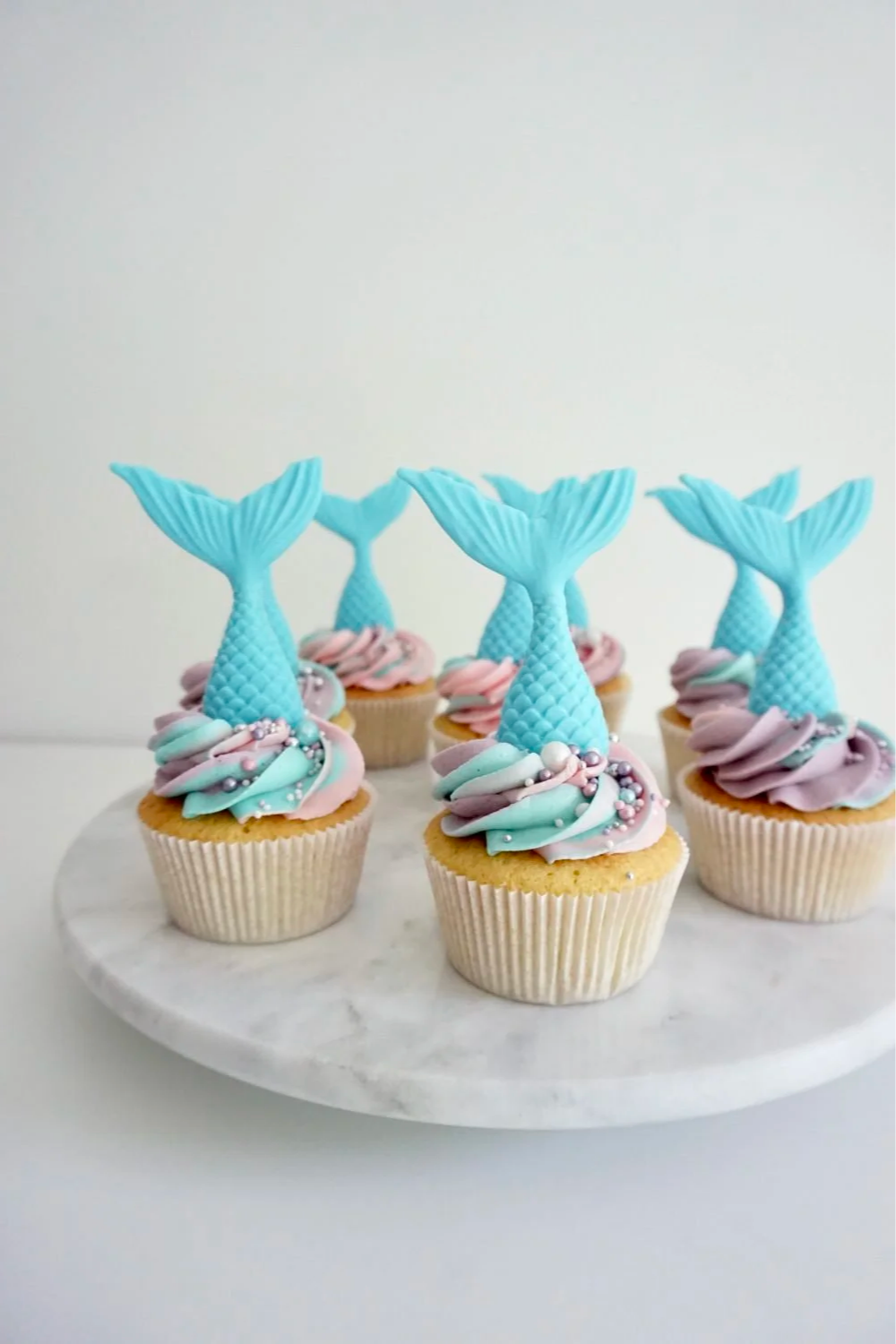 Cupcakes with pastel-colored frosting and mermaid tail decorations on top, arranged on a white marble cake stand.