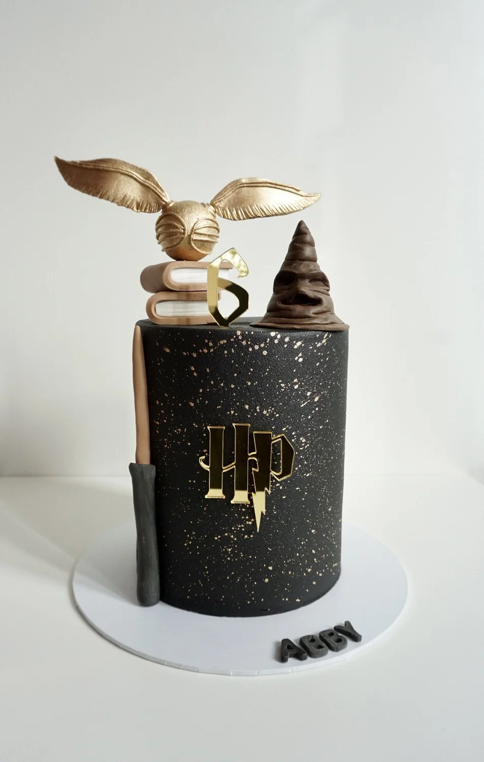 A Harry Potter themed birthday cake with a black base decorated with gold splatters. On top, there is a golden owl, a book, a golden key, and a chocolate broomstick. The cake has a Harry Potter logo and the word 'AOSY' on the base.