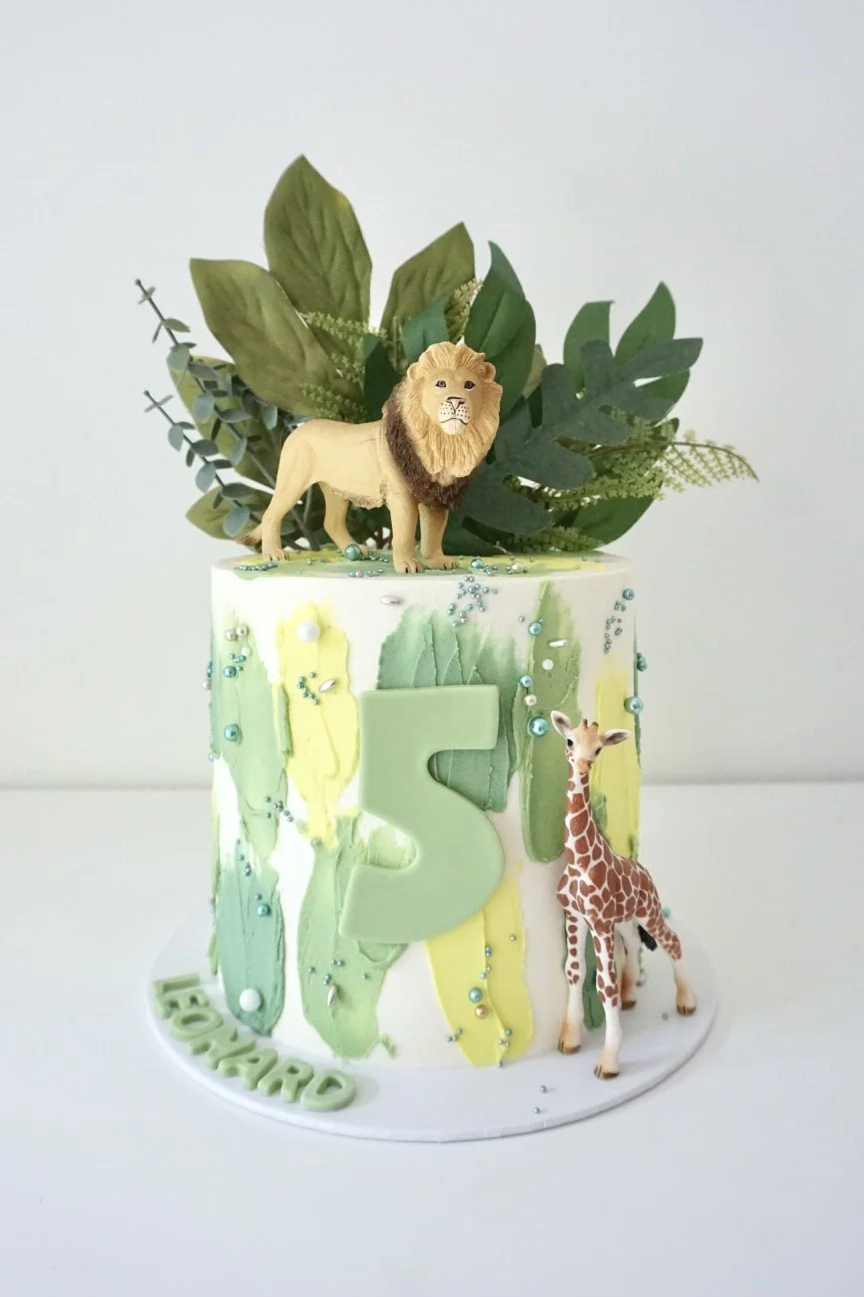 A birthday cake decorated with a green and white marbled pattern, with a blue number four on the front. The cake has artificial plants and a toy dinosaur on top, themed for a child's birthday.