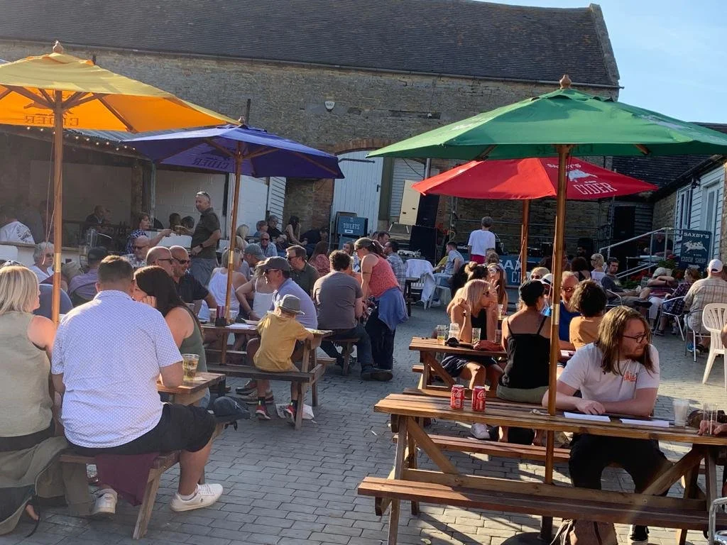 Outdoor gathering with people sitting at picnic tables under colorful umbrellas, enjoying food and drinks, in front of a stone building with a stage set up for a performance.