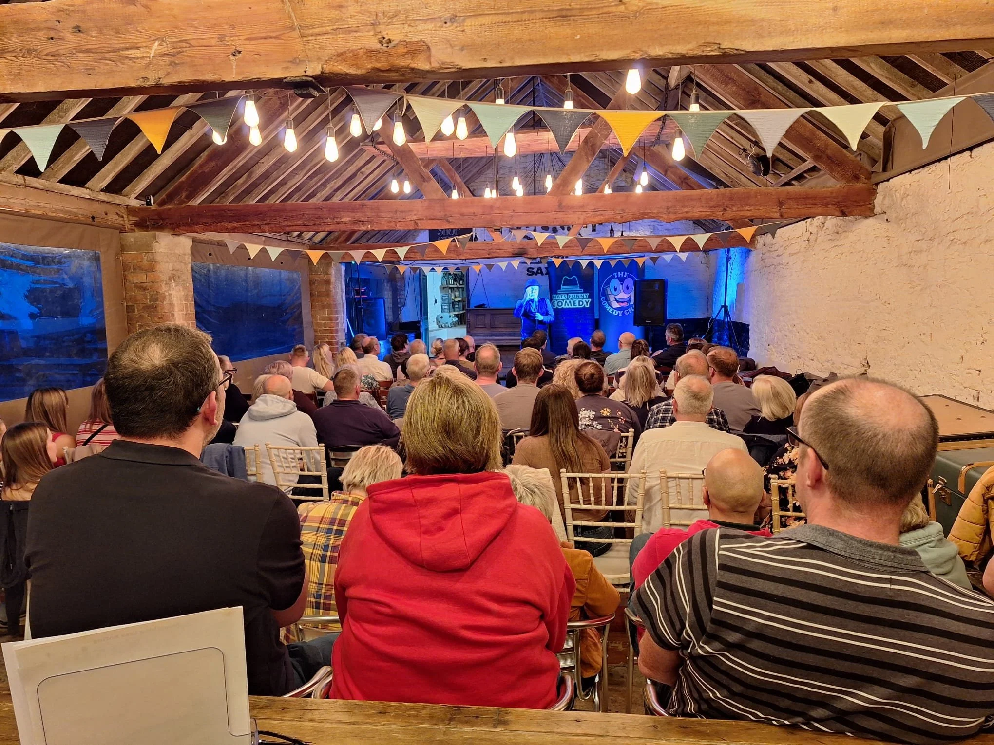 Audience watching a comedian perform on stage in a rustic venue decorated with string lights and pennant banners.