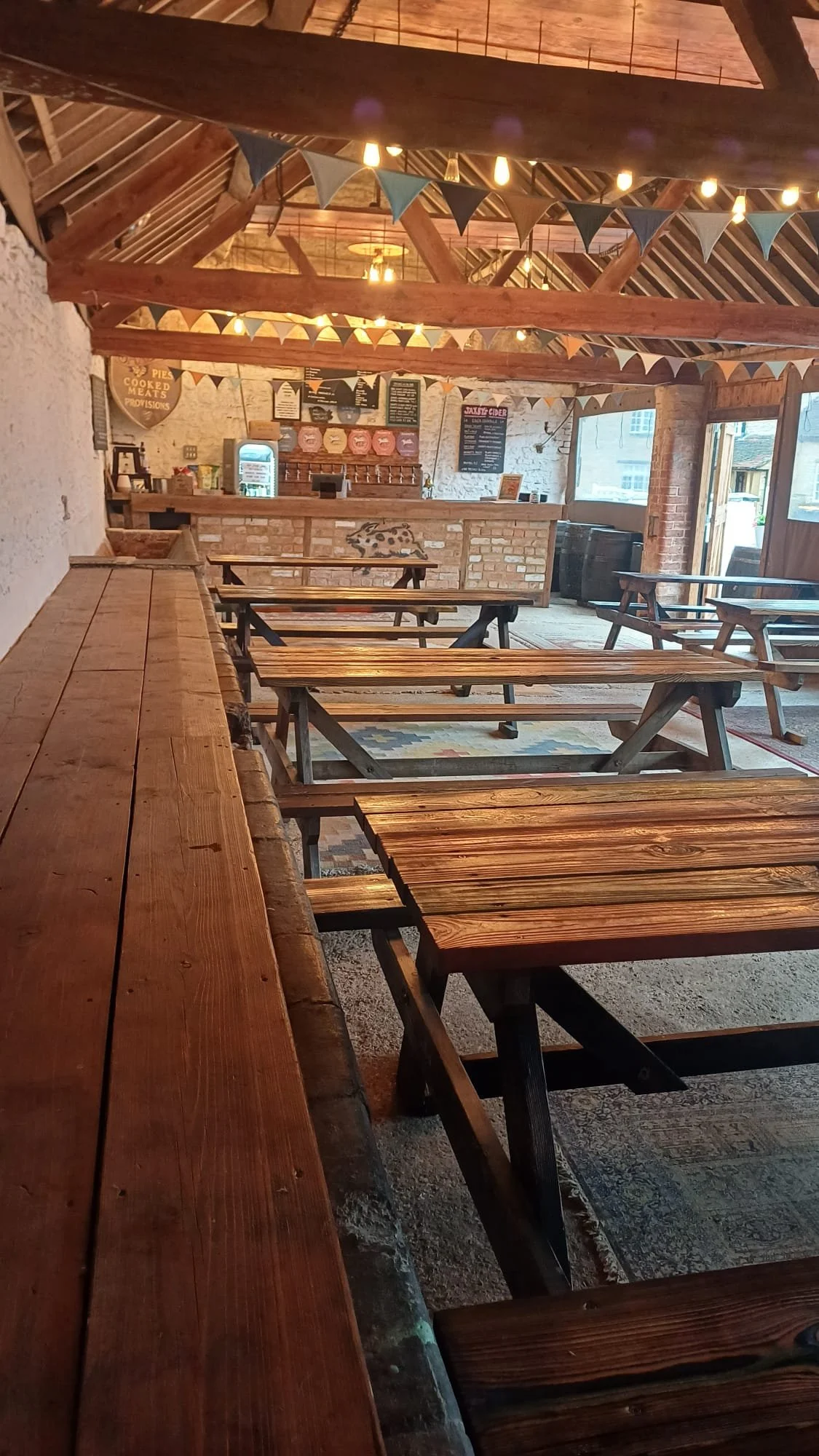 Interior of a rustic-style restaurant or pub with wooden benches and tables, a high ceiling with exposed wooden beams and string lights, and a brick counter area with menu boards and a small refrigerator.