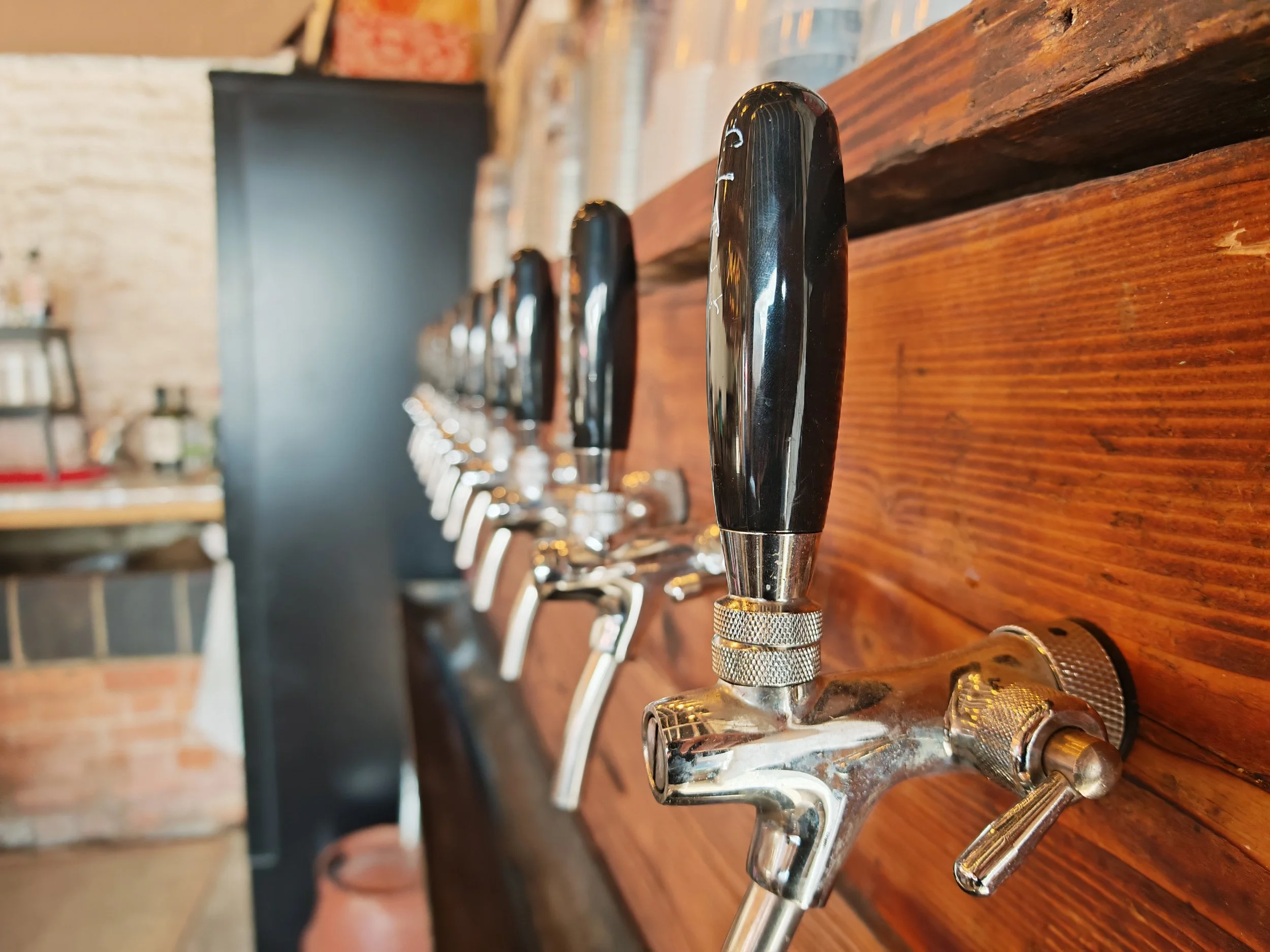Close-up of beer taps with black handles mounted on a wooden bar, with a background of exposed brick wall and shelves.