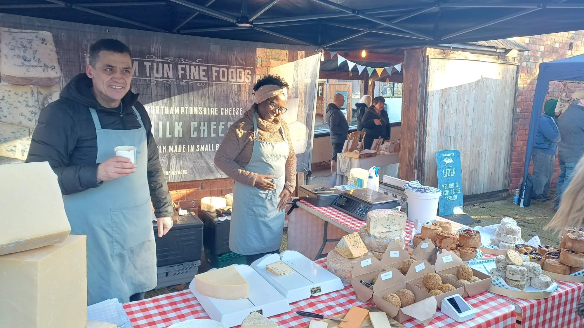 Market stall selling cheese and cheese-based snacks, with two vendors behind the table, one holding a drink and the other standing with hands in pockets, under a canopy with string lights and a sign in the background.
