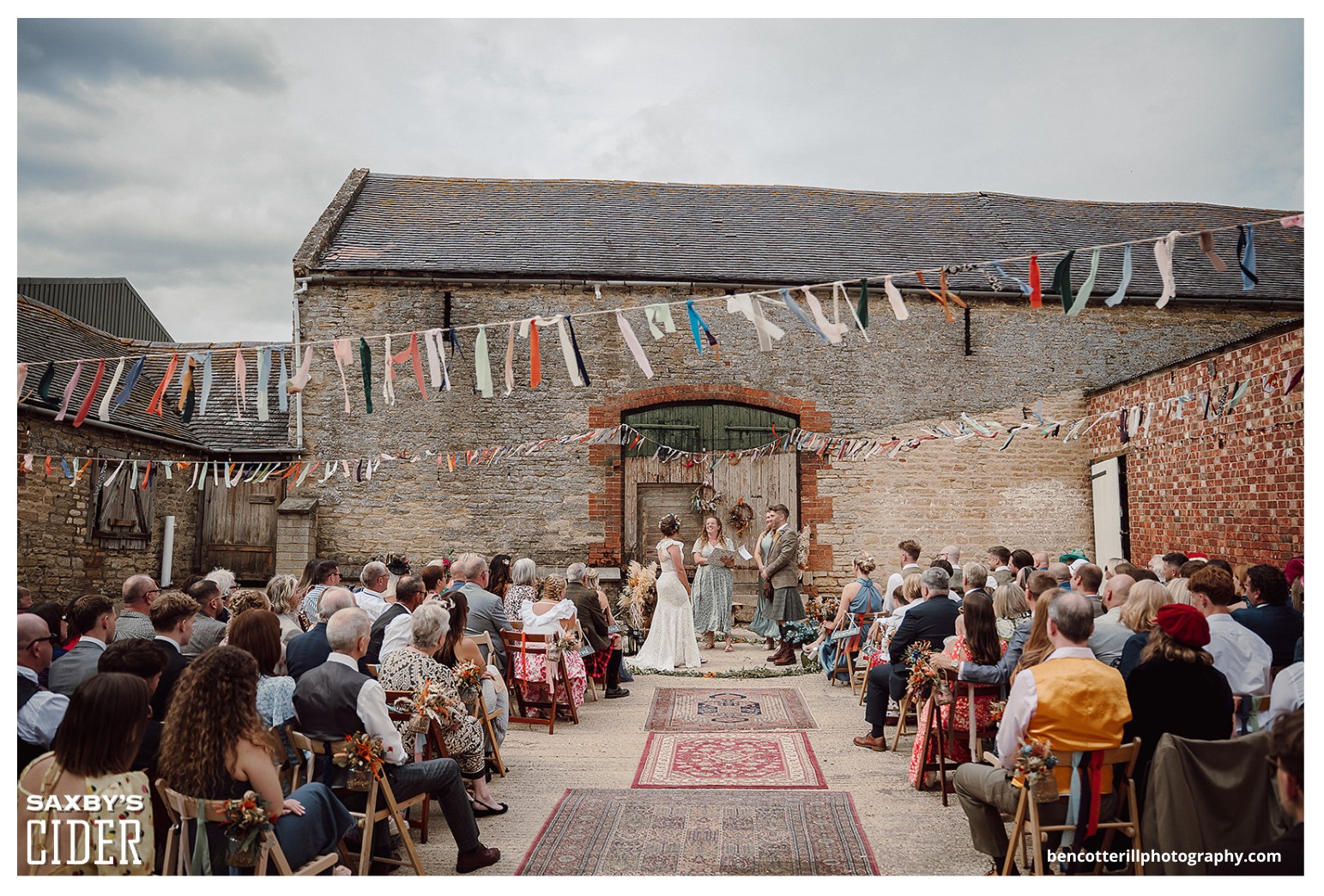 Outdoor wedding ceremony in a rustic barn setting with guests seated on wooden chairs, facing the couple and officiant at the front, decorated with colorful ribbons and floral accents, against an old brick and stone barn wall.
