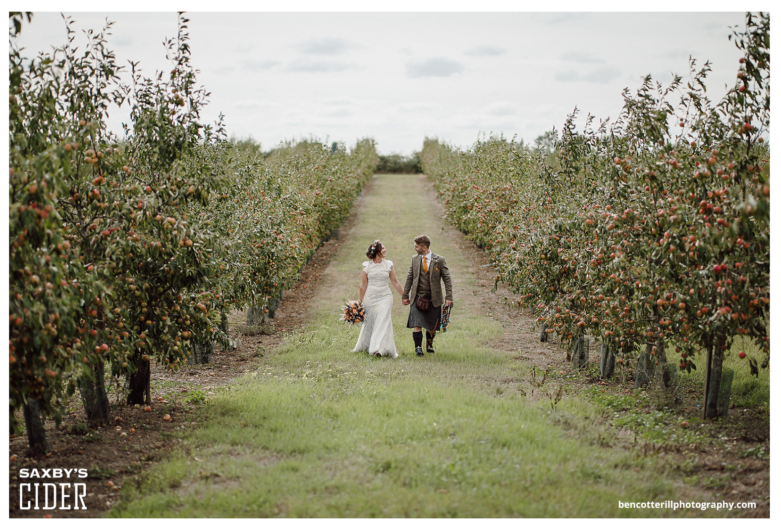 A bride and groom holding hands walking through an apple orchard on a cloudy day, with the bride holding a bouquet of flowers.