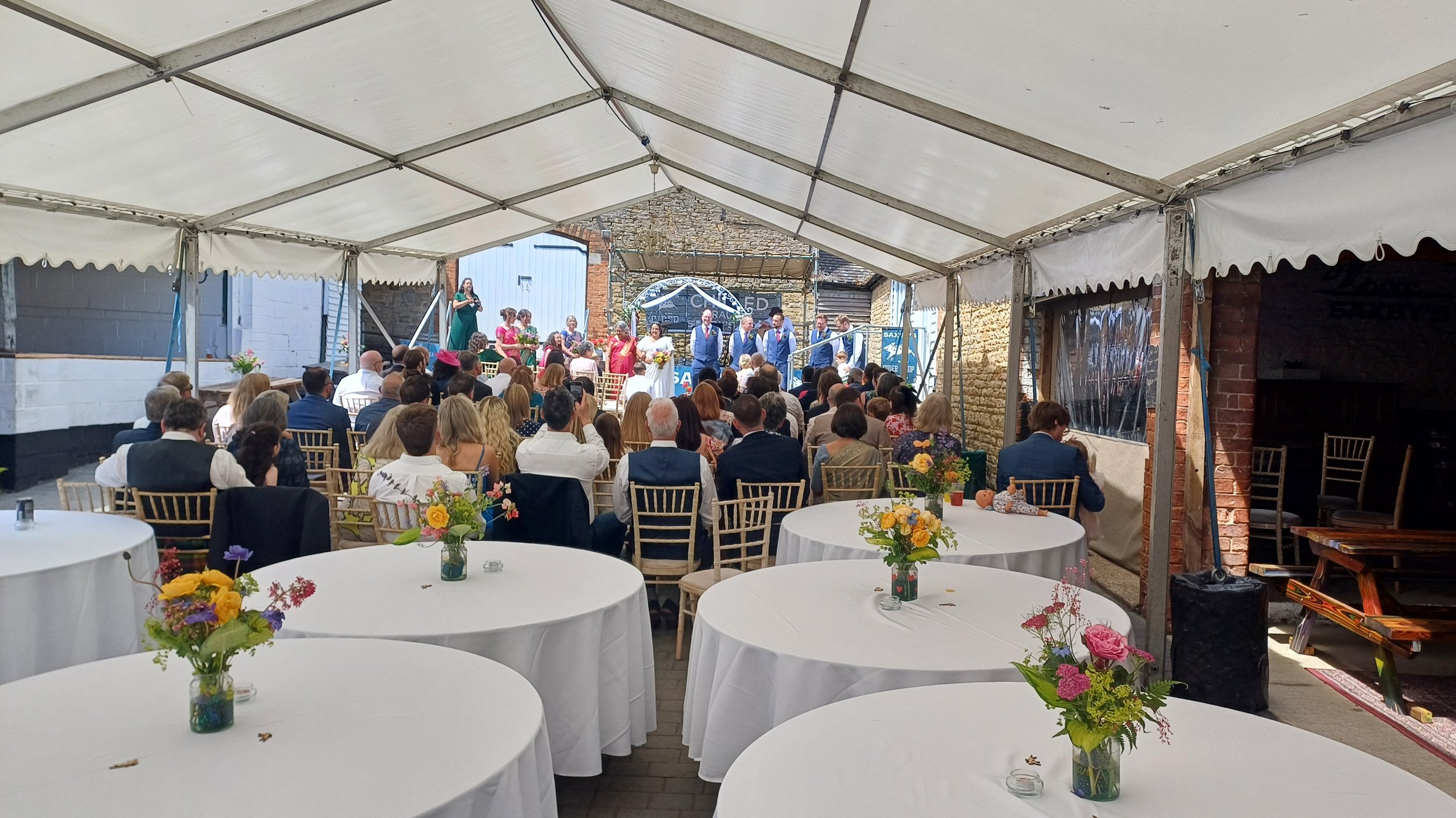 An outdoor wedding under a large white tent with guests seated at round tables decorated with vases of colorful flowers. On the stage at the front, a bridal party is present, with the bride in a white dress and the groom in a gray suit. Other people 