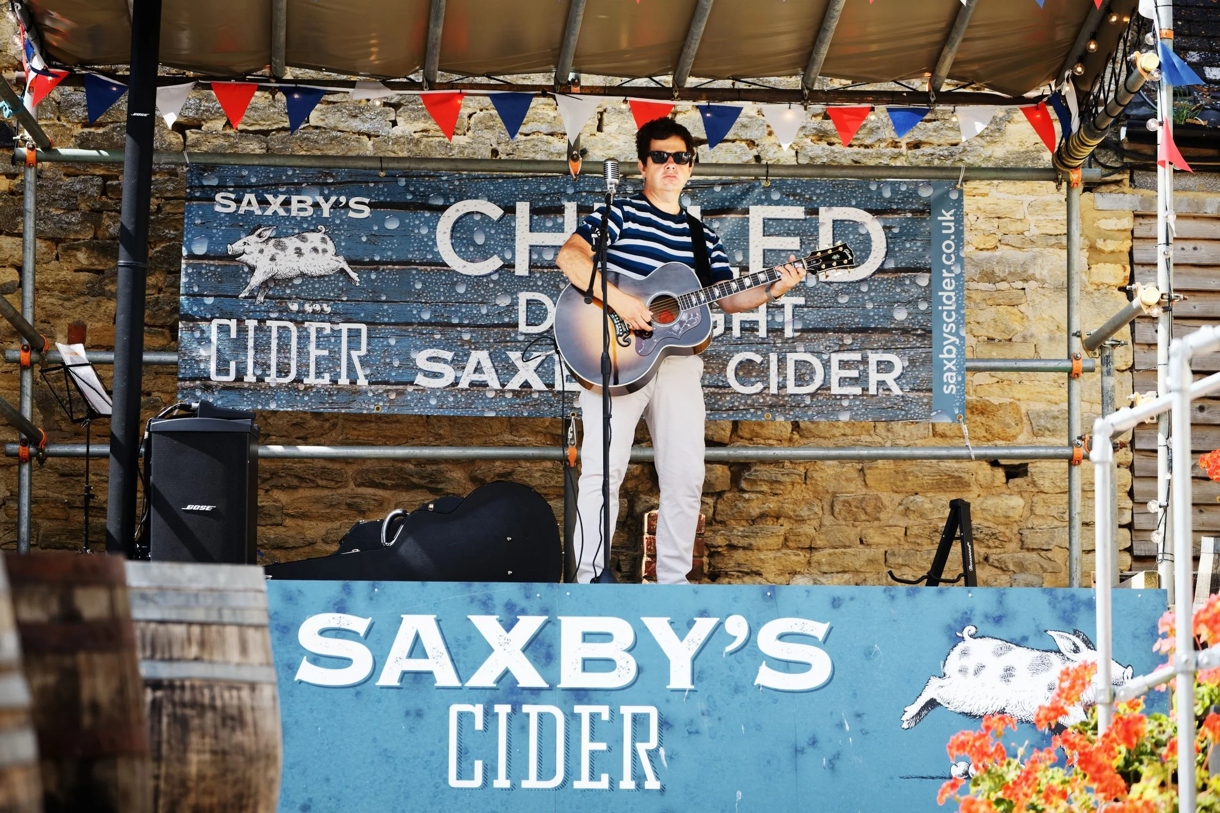 A musician playing an acoustic guitar and singing into a microphone on a small outdoor stage, with a backdrop advertising Saxby's Cider, decorated with red, white, and blue banners.