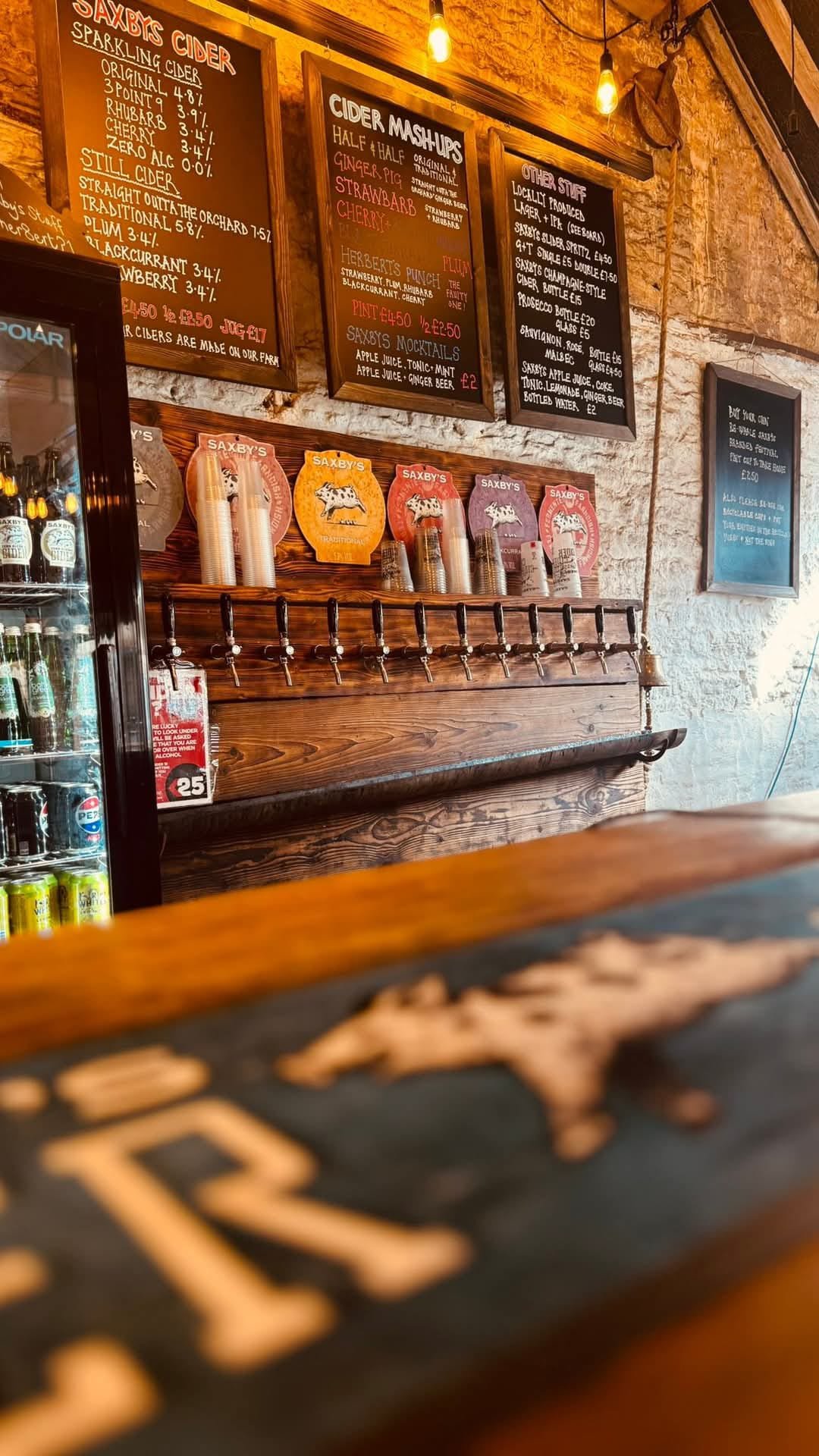 View of a pub or bar counter with tap handles, drink menus on blackboards above, and a refrigerator with bottled drinks to the left.