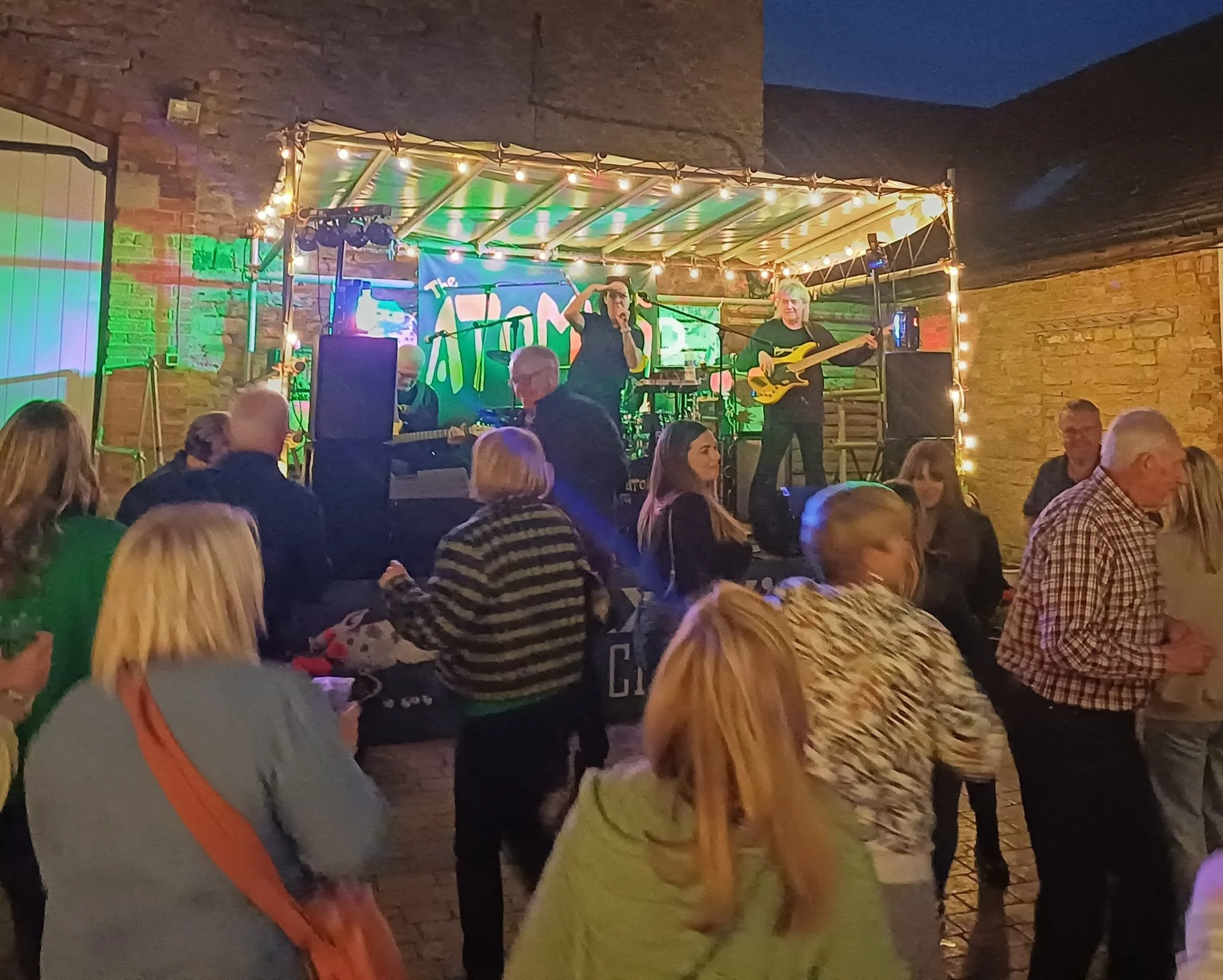 Live music performance on a stage with a band, playing instruments and singer, at an indoor or outdoor venue during evening, with audience dancing and socializing.