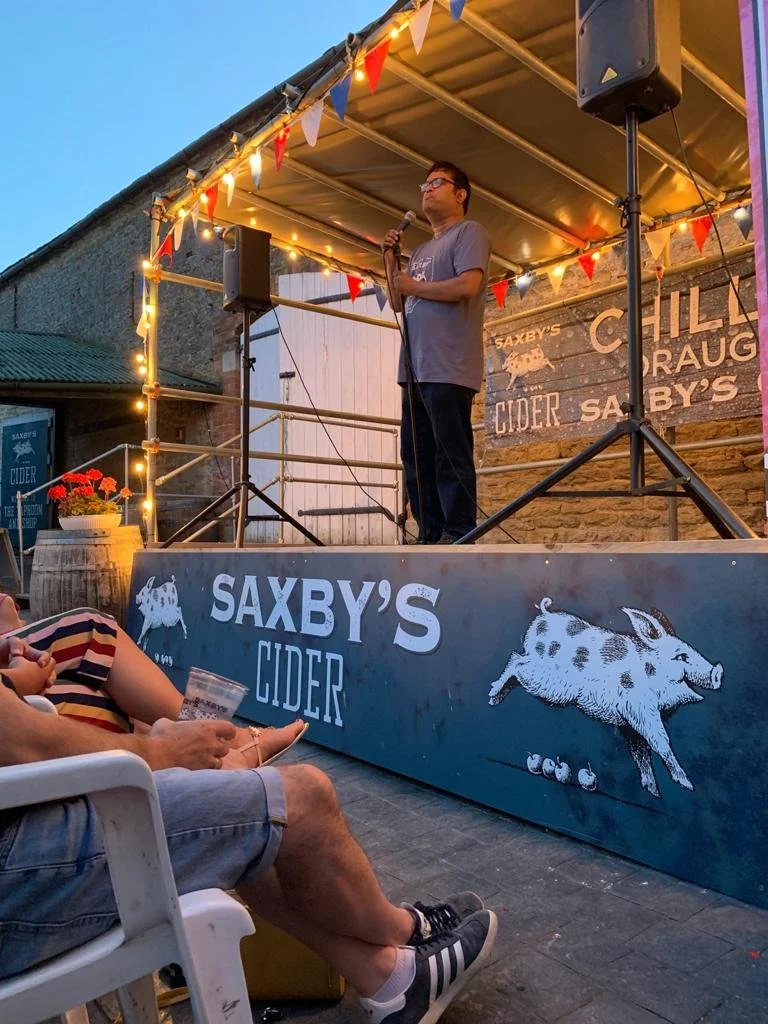 Man on stage with microphone at outdoor venue, decorated with string lights and red, white, and blue pennant banners, side view of audience members seated and using phones or drinks, background features sign for Saxby's Cider with pig and boar illust
