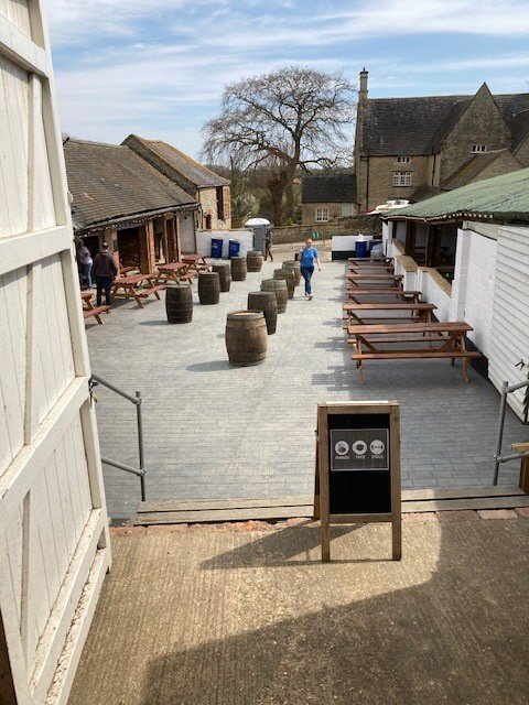Outdoor patio area of a pub or restaurant with wooden benches, tables, barrels, and trash bins, with a few people walking around.