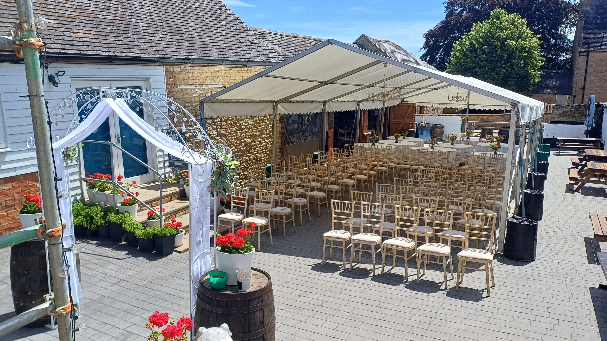 Outdoor event setup with rows of chairs under a white canopy, surrounded by potted red and green plants, in a paved courtyard with picnic tables and a building in the background.
