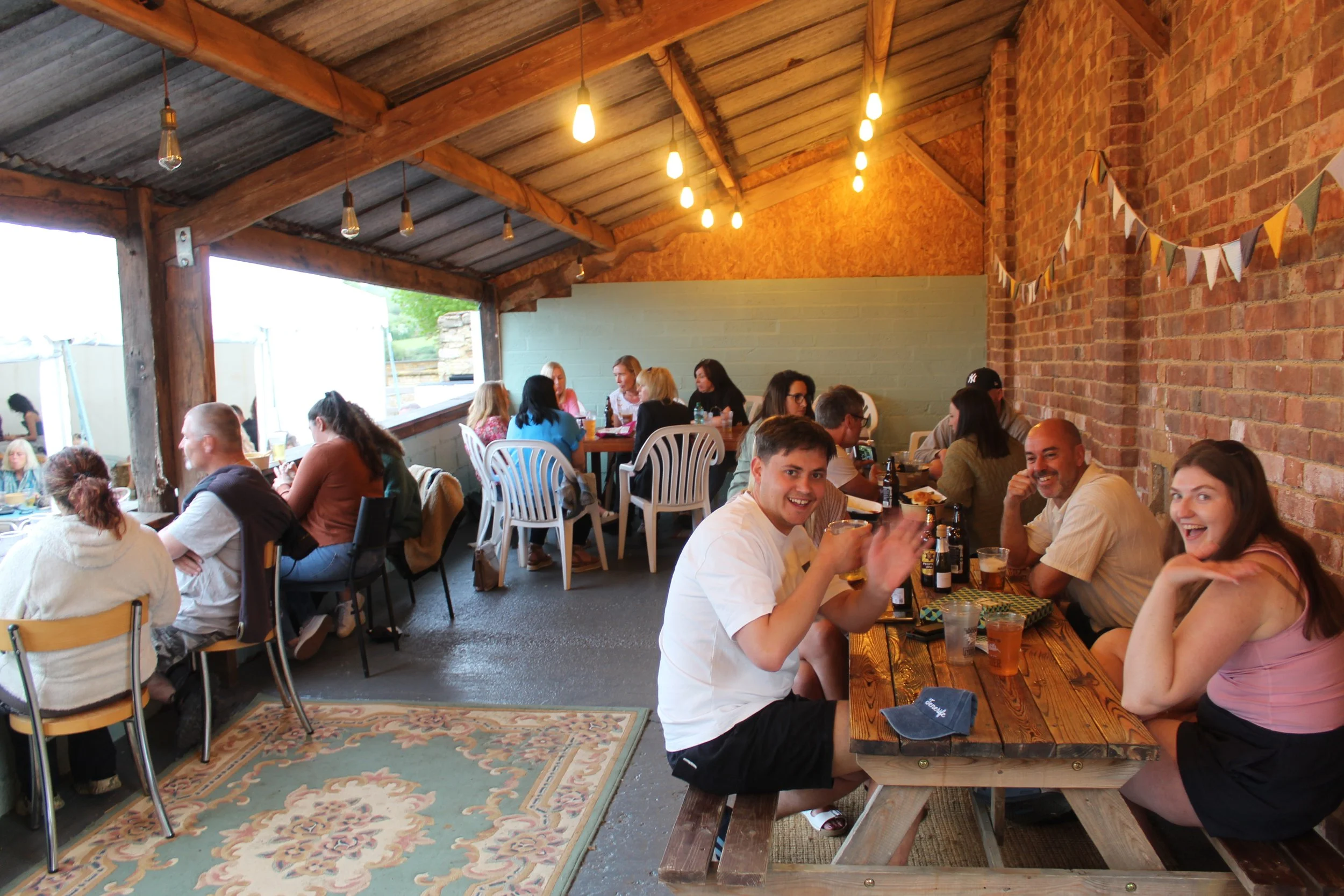 People enjoying a casual outdoor gathering at a covered patio with exposed brick walls, string lights, and a rug, sitting at tables with drinks and food, smiling and socializing.