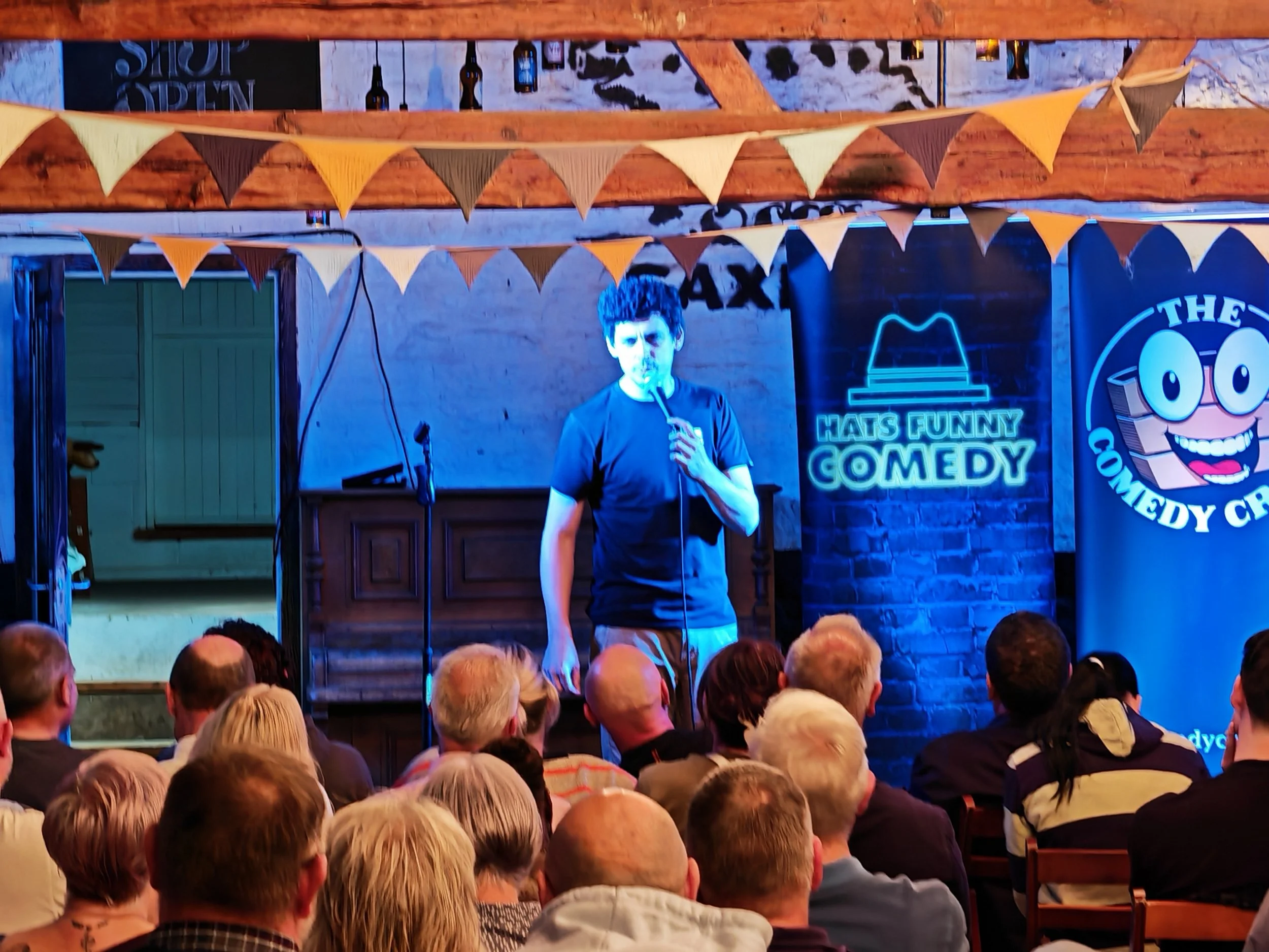 Comedian performing on stage at a comedy club with an audience watching.