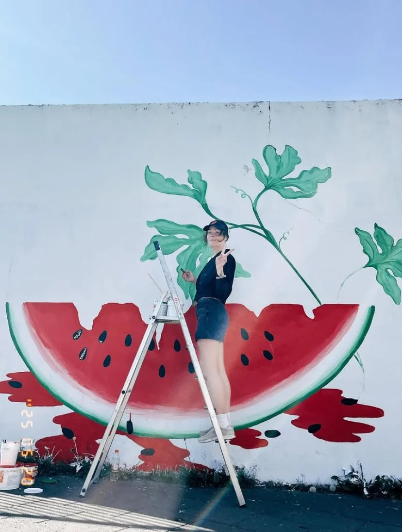 A woman standing on a ladder in front of a wall mural of a watermelon slice. The woman is making a peace sign and appears to be painting or working on the mural.