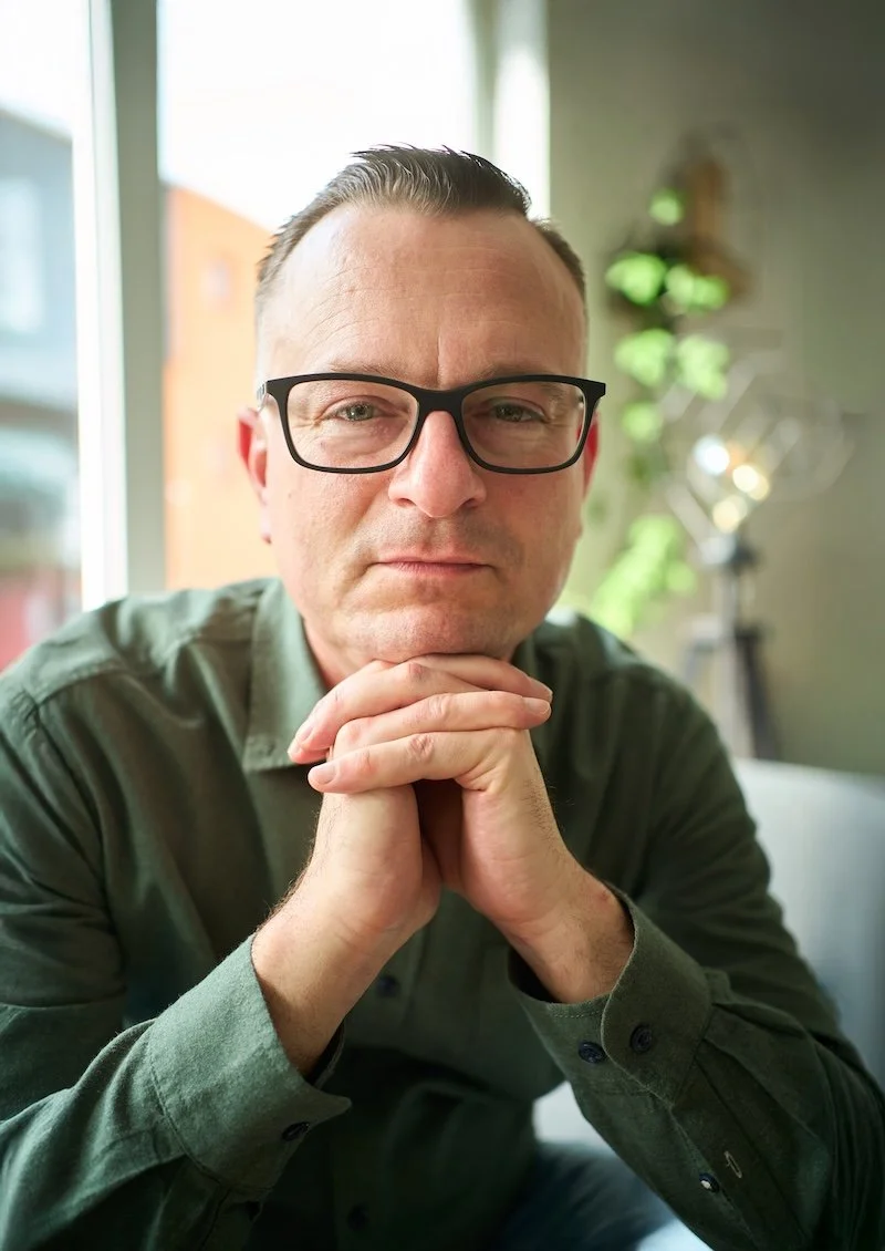 Rico Otterbach sitzt drinnen an einem Tisch, trägt ein grünliches Hemd und schaut in die Kamera, wobei er die Hände unter dem Kinn verschränkt.