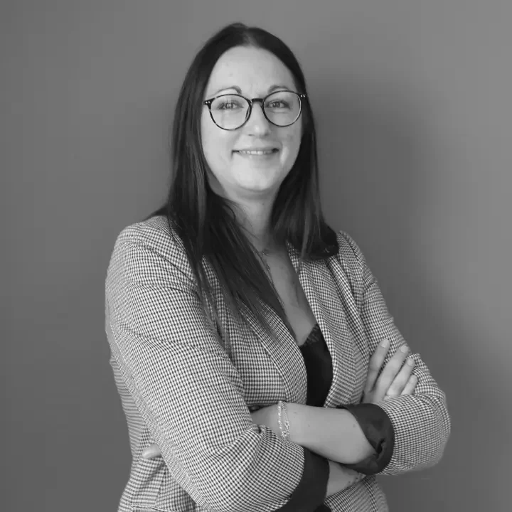 une femme souriante avec des lunettes, portant une veste à motifs et un haut noir, croisant les bras, posant contre un mur gris.