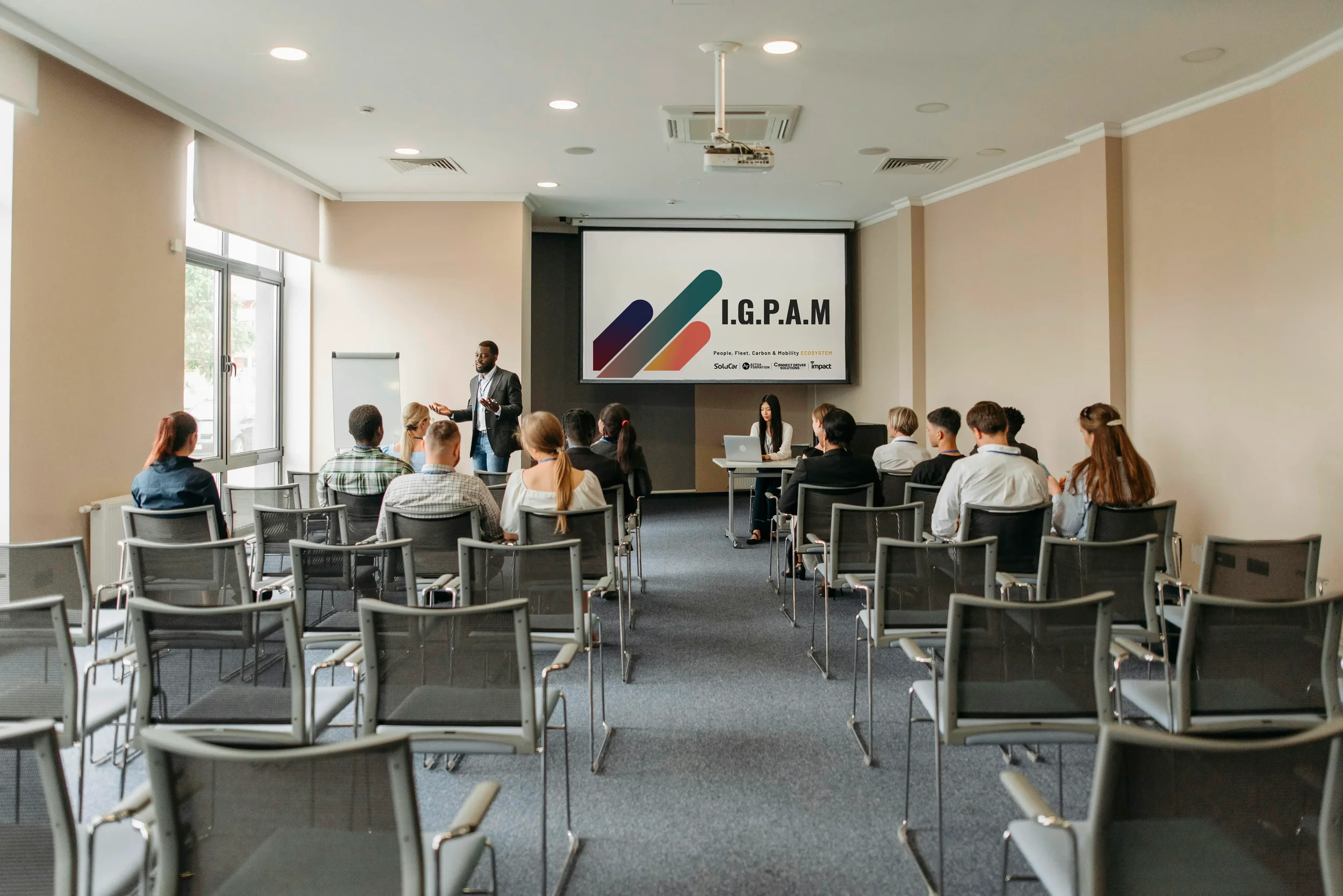 Une conférence se déroulant dans une salle avec des chaises disposées en rangées. Un homme en costume donne une présentation devant un grand écran affichant 'I.G.P.A.M'. Une femme est assise à une table avec un ordinateur portable, face à l'audience.