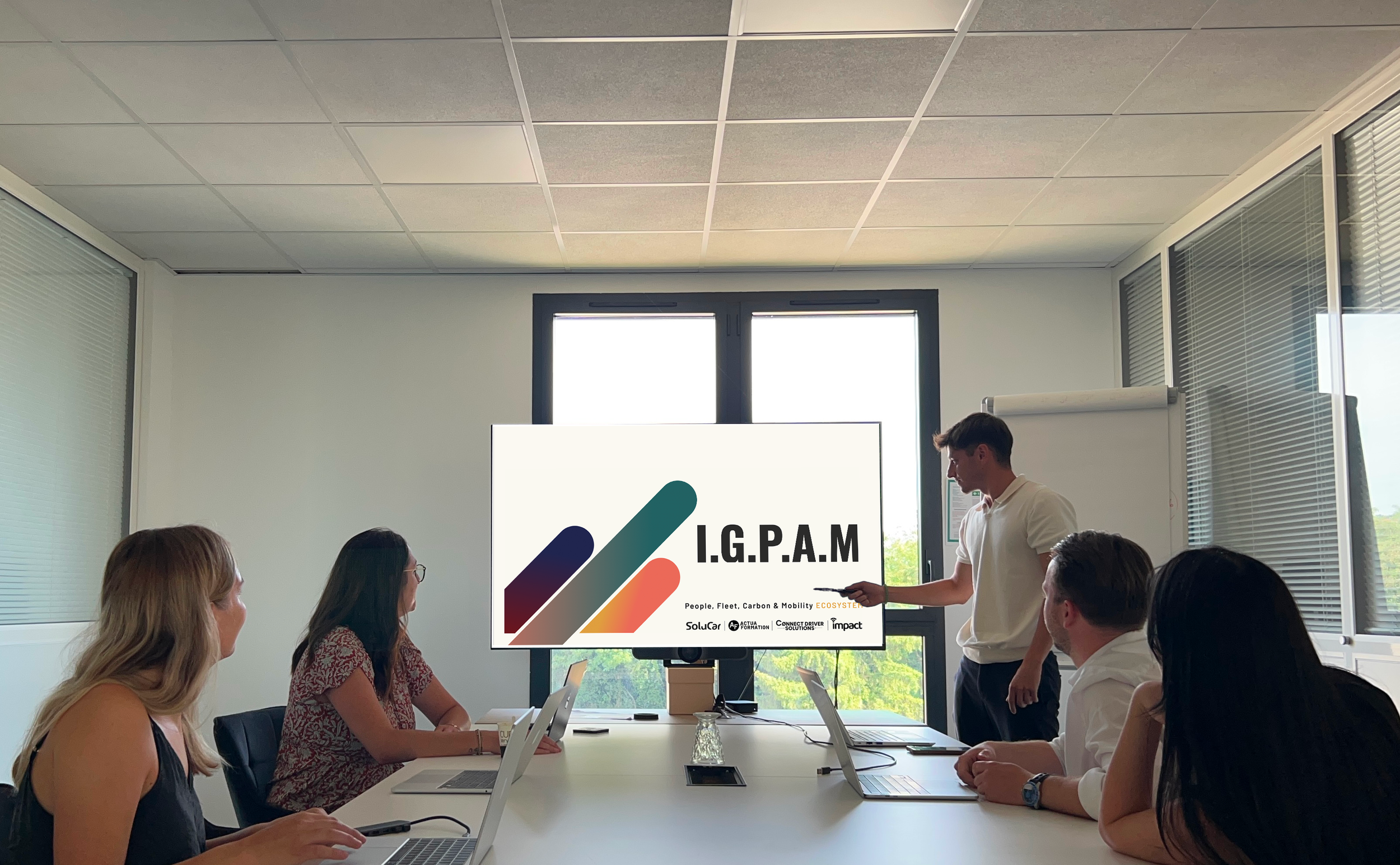 Une réunion de groupe dans une salle de conférence, avec un présentateur qui montre un écran affichant "I.G.P.A.M" et un graphique. La salle a de grandes fenêtres laissant entrer de la lumière, et les participants utilisent des ordinateurs portables.