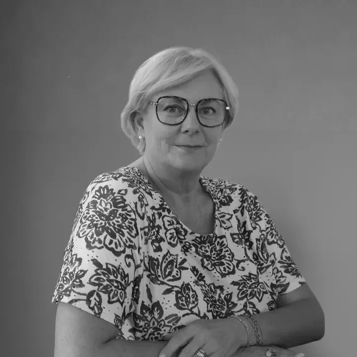 Portrait d'une femme âgée portant des lunettes, avec une chemise à motif floral, les bras croisés, sur un fond uni.