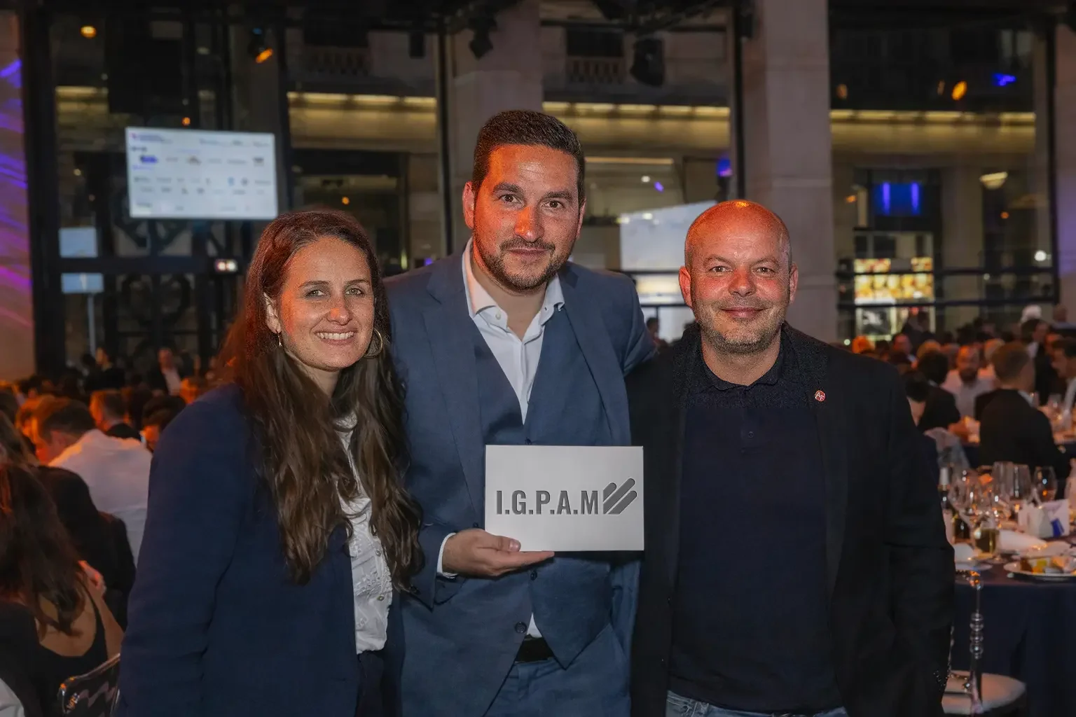 Trois personnes posant ensemble lors d'un événement formel, deux hommes et une femme, avec une salle remplie de tables et de convives en arrière-plan.