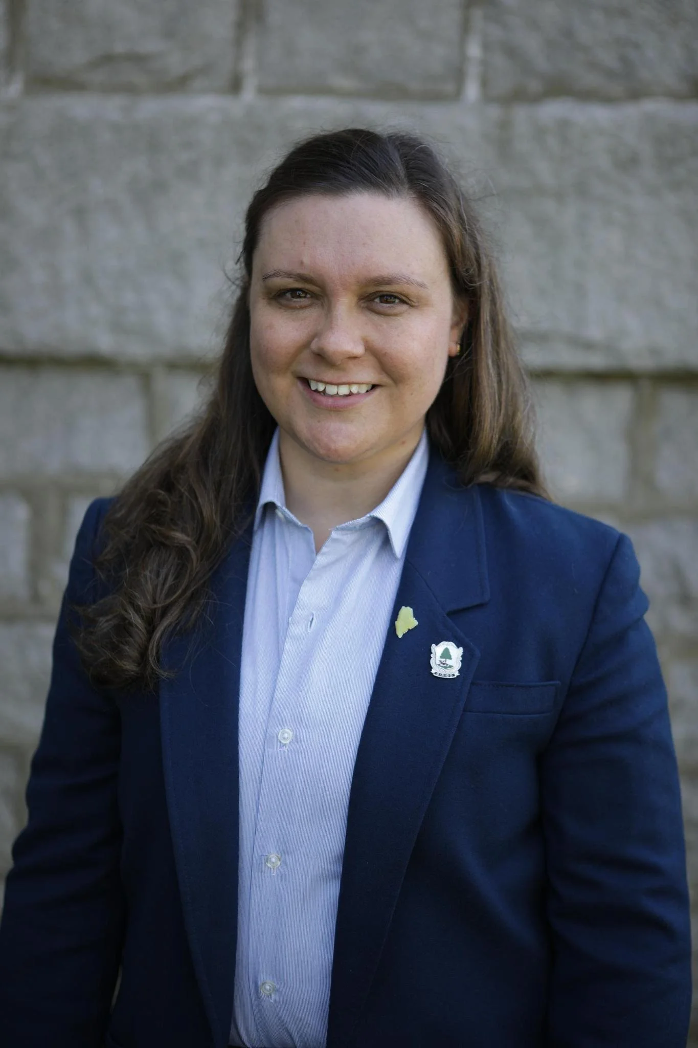 Andrea LaFlamme, a US Senate candidate for Maine,  is smiling towards the camera. She is standing outside against a wall backdrop and is wearing a blue blazer jacket.