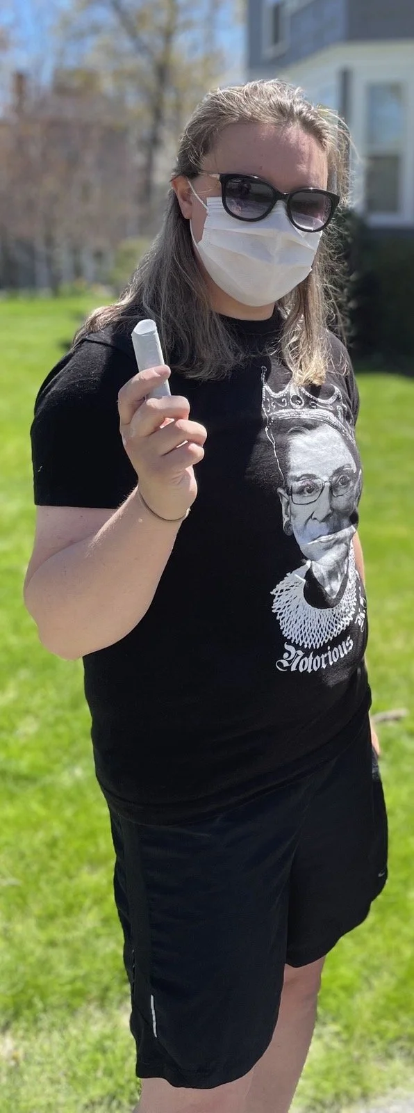 Andrea LaFlamme is standing outside Susan Collins house. She is wearing a face mask and sunglasses and is holding up a piece of chalk.