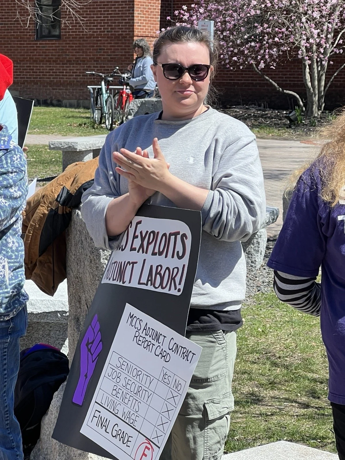 Andrea LaFlamme is outside at a protest gathering. She is wearing sunglasses and a gray sweatshirt, and holding a protest sign (sign text is not fully viewable). She is clapping her hands in response to a speaker.